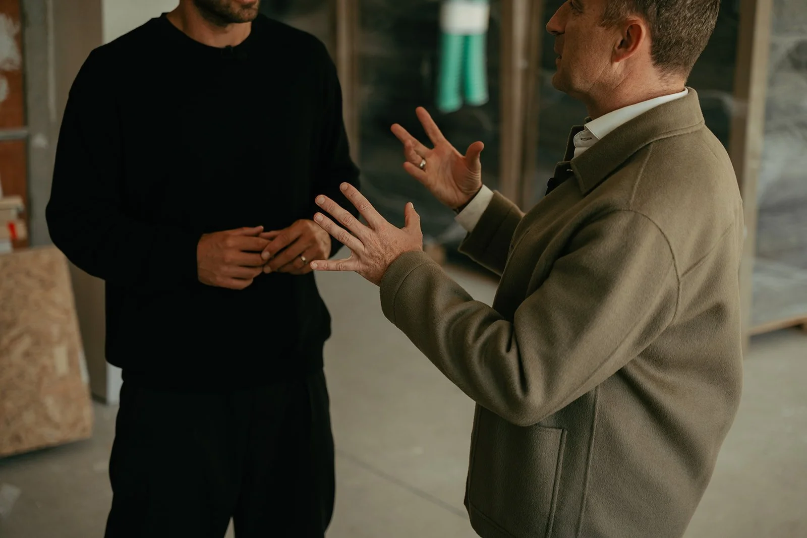 Two men are engaged in conversation indoors, one is gesturing with his hands while the other listens with his hands clasped.