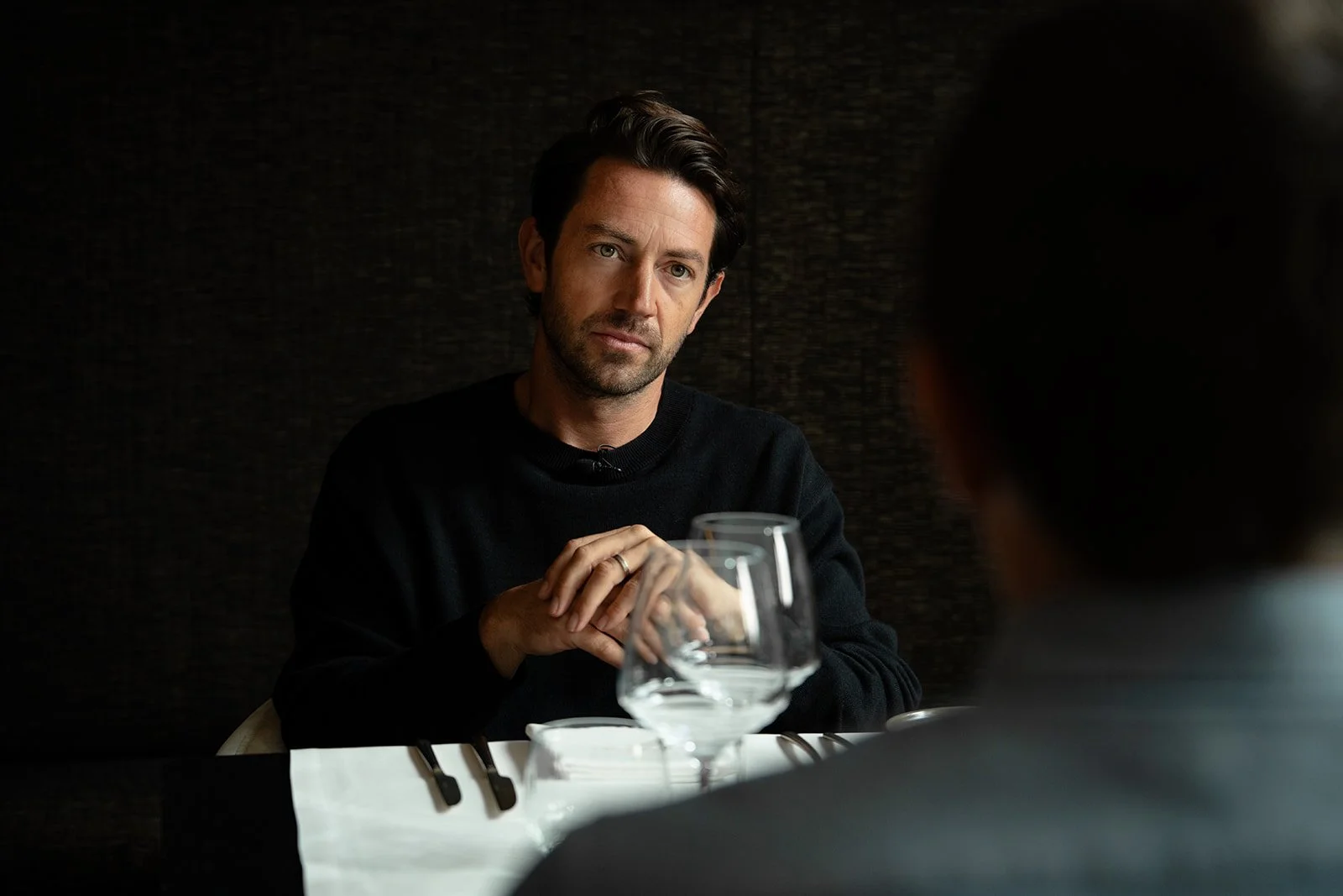 Man with dark hair and beard wearing a black sweater, sitting at a table in a restaurant with a dark textured wall, engaged in conversation with another person whose back view is visible.