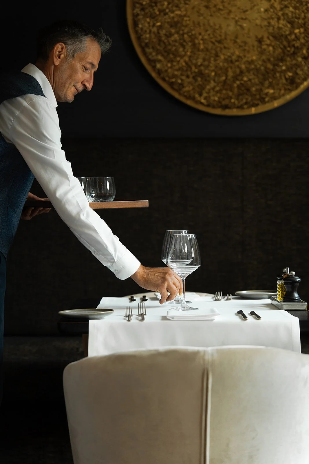 A man in formal attire setting a table with glasses and cutlery in a dimly lit restaurant.