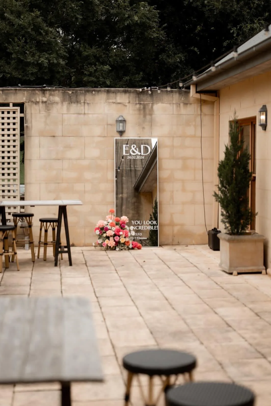 Wedding venue outdoor patio with a large mirror sign that reads 'E & D 24.02.2024' and 'You look incredible!'. Decorated with a bouquet of pink and white flowers placed at the base of the mirror, and green potted plants on the right. There are small tables and stools on the left, string lights overhead, and wall-mounted lanterns. The patio is made of stone tiles, and there is a beige brick wall with a lantern in the background.