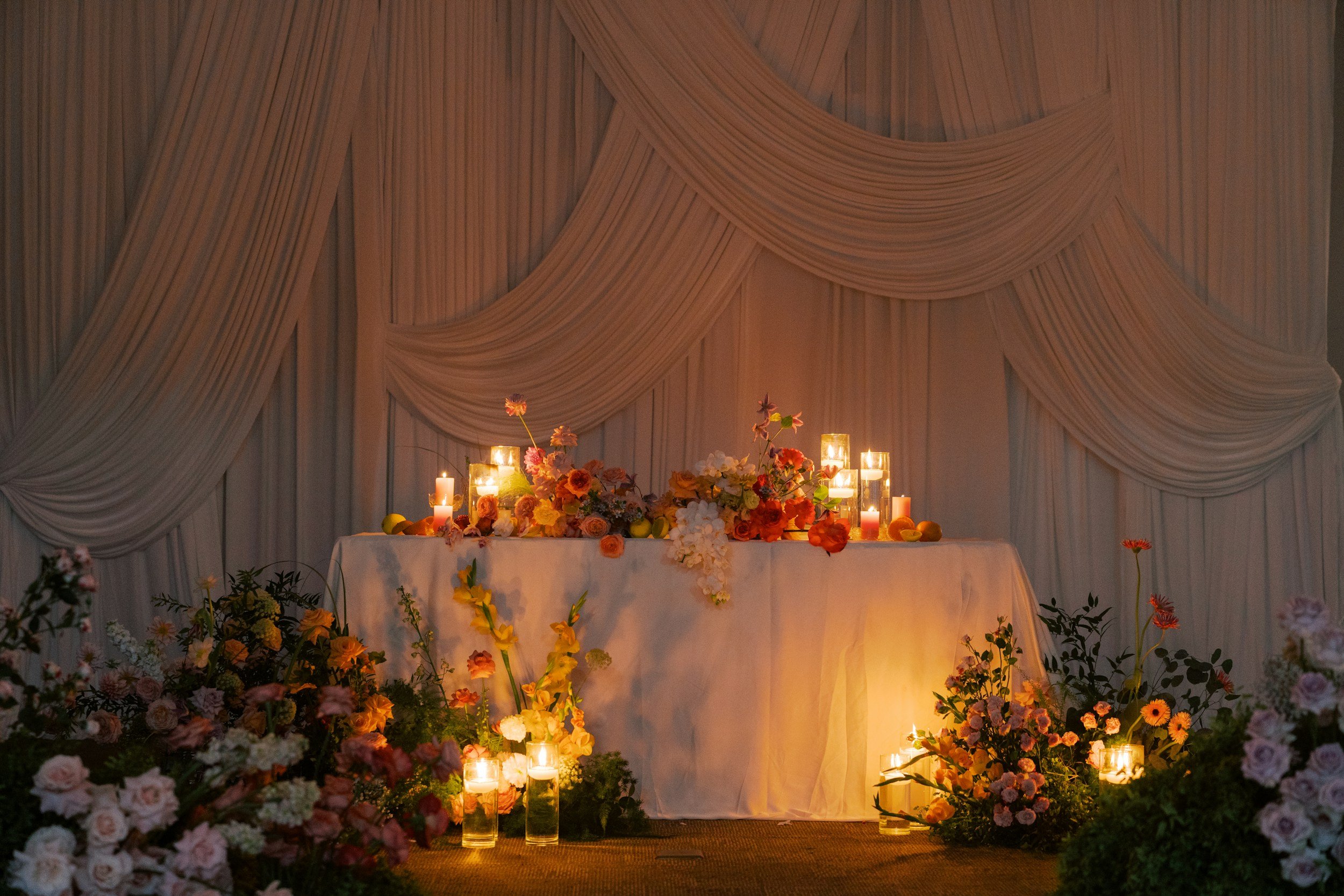 A decorated table with candles and flowers in front of a beige draped curtain, likely for a formal event or celebration.
