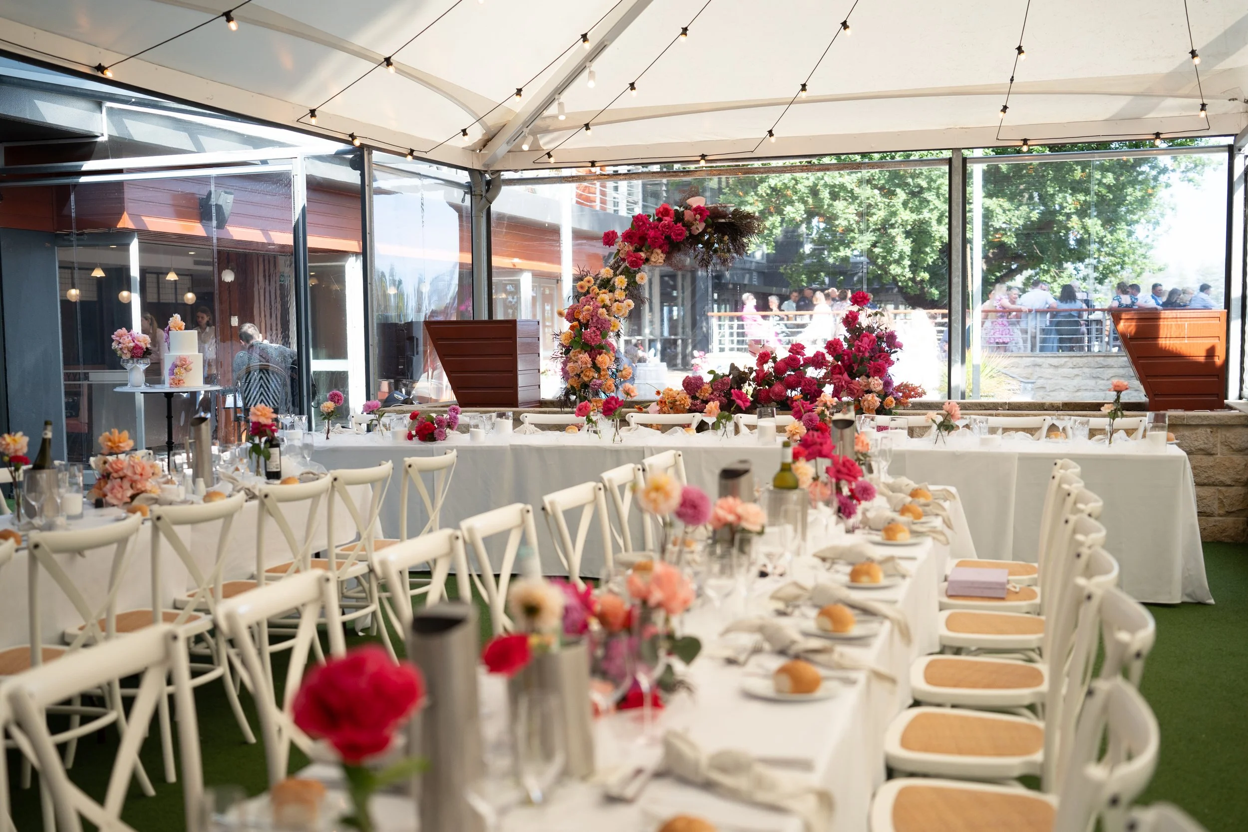 Indoor event space decorated for a celebration with round tables covered in white tablecloths, pink and orange flowers, candles, and place settings, featuring a floral arrangement with pink, orange, and yellow flowers and a cake on a stand. The space has large glass windows with views of trees and people outside, and string lights hanging from the ceiling.