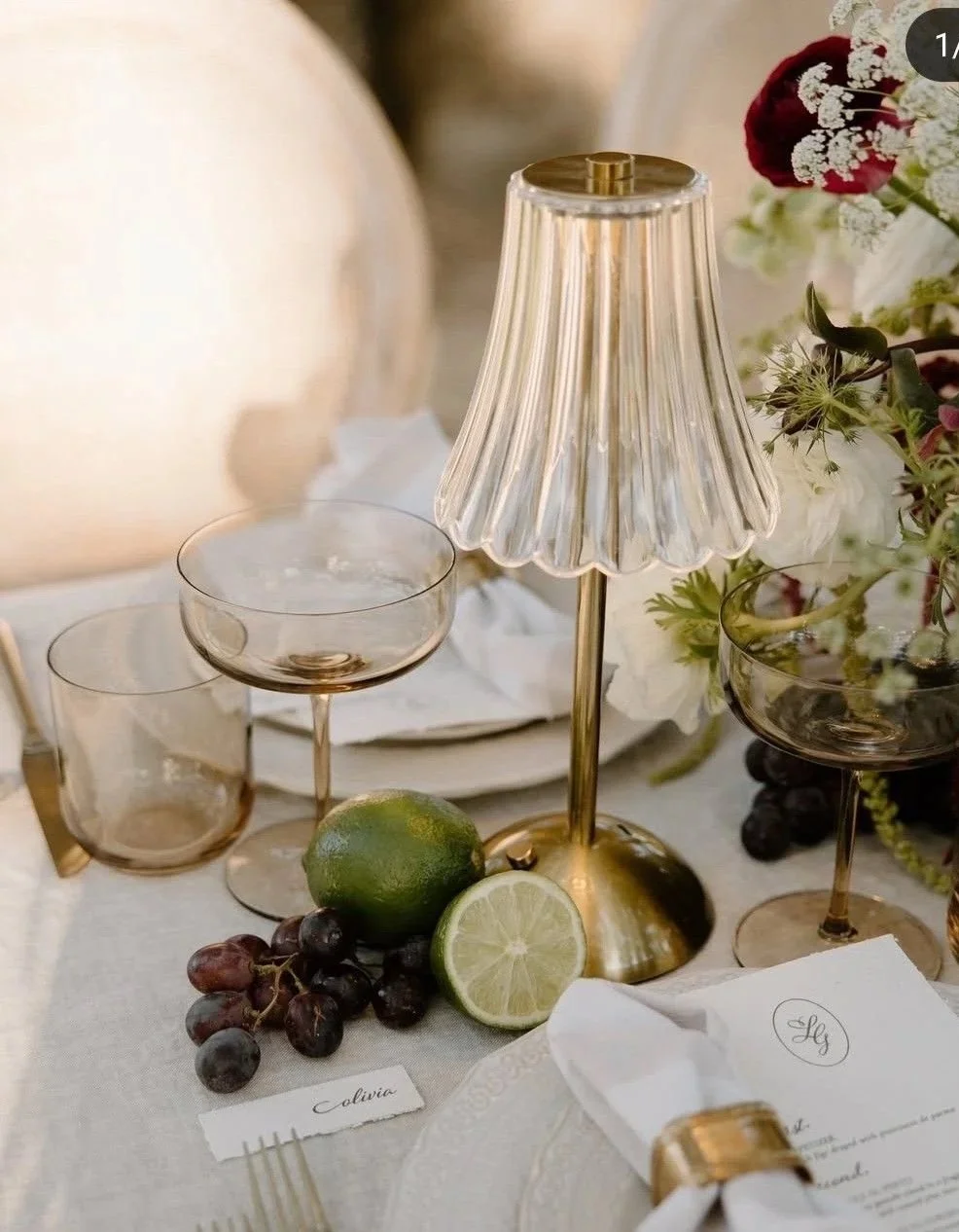 Elegant table setting with a gold and glass lamp, grapes, lime, and floral arrangements.