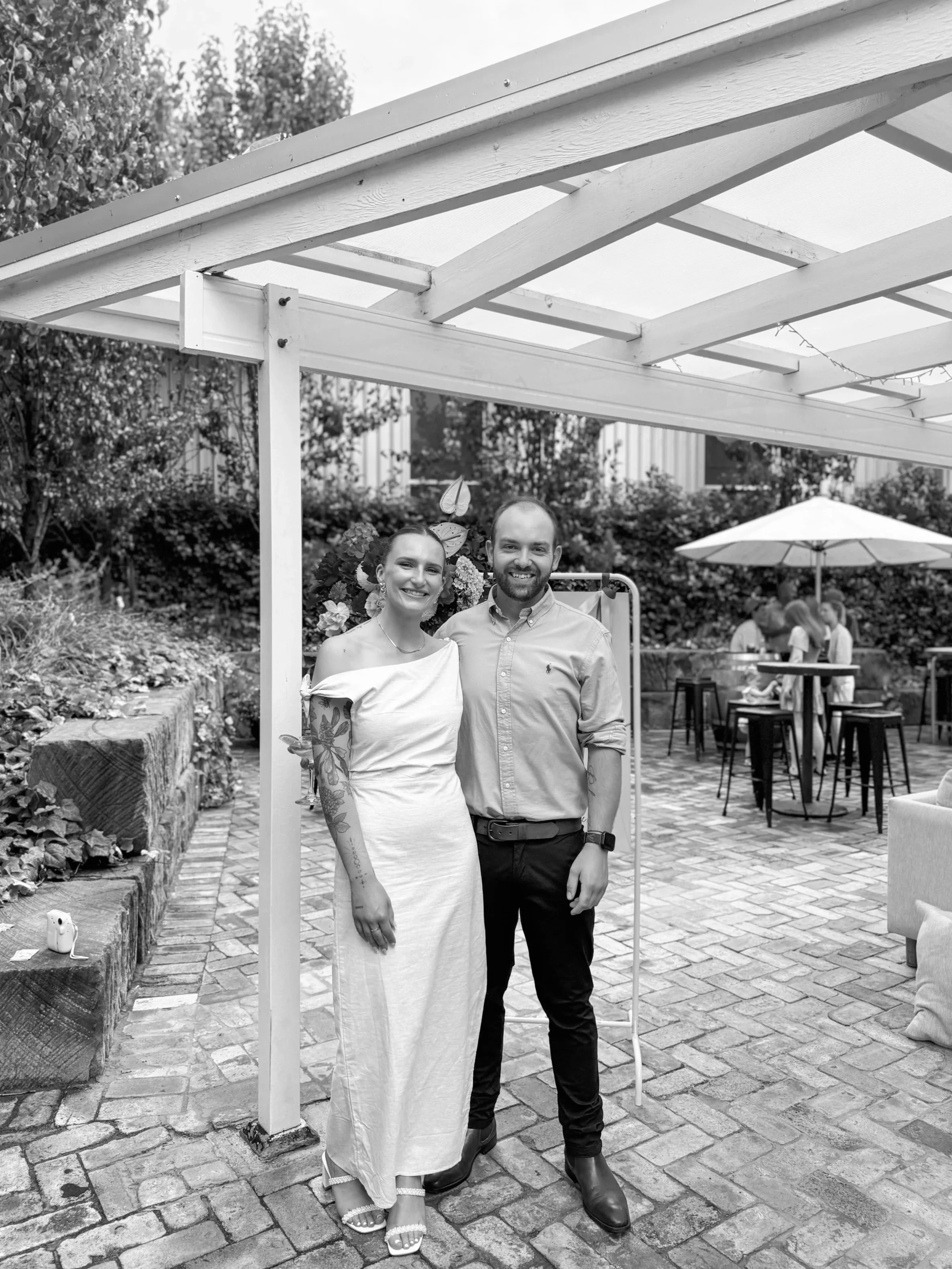A woman and a man standing close together, smiling at the camera, outdoors at a social gathering. The woman is in a white off-shoulder dress with tattooed arm, and the man is wearing a light-colored button-up shirt and dark pants. They are under a gazebo with a brick patio, with people and table with umbrella in the background.