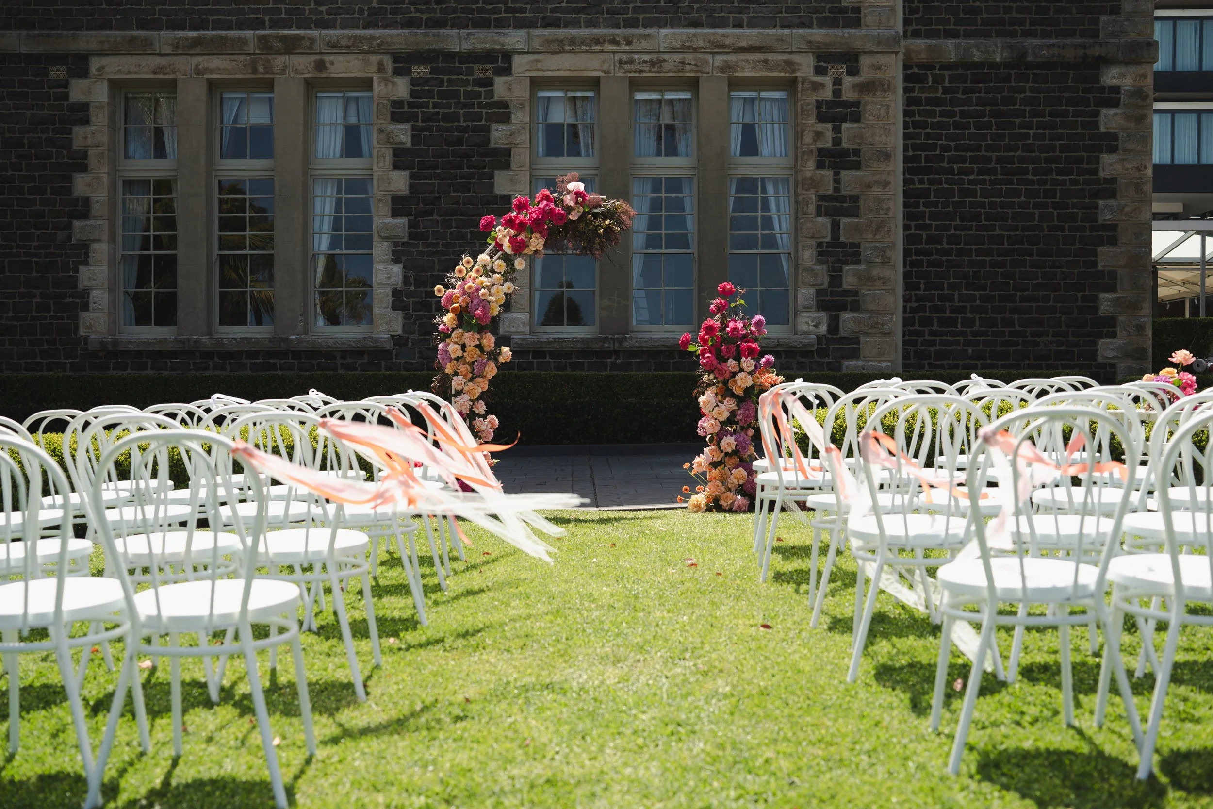 Outdoor wedding ceremony setup with white chairs, floral arch, and a brick building in the background.
