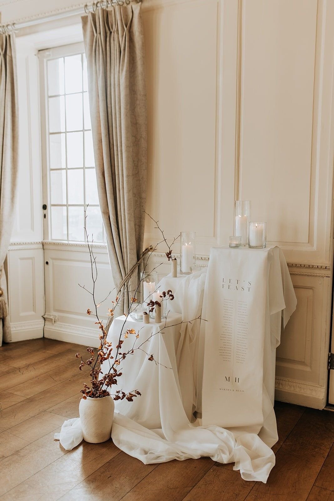 Decorative table with candles, a printed cloth, a bouquet of dried branches in a white vase, set against cream-colored walls and window with beige curtains.