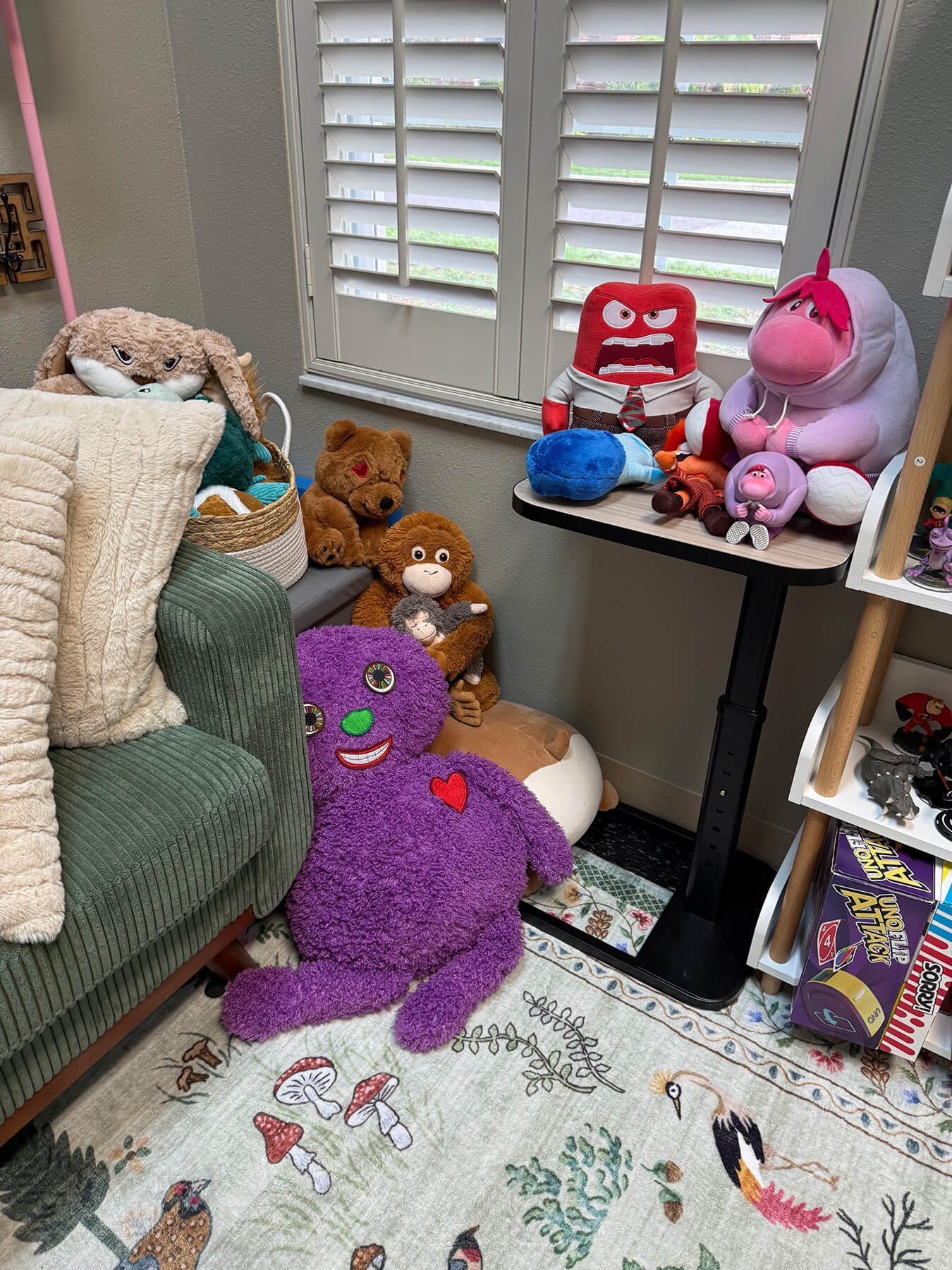 Collection of plush toys and stuffed animals arranged in a room with a patterned rug, window with blinds, and a small table.