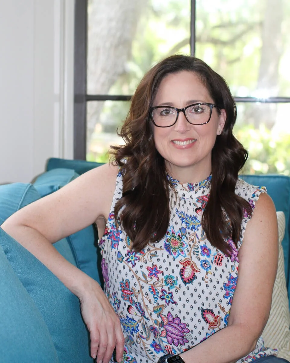 A counselor woman with glasses and long, wavy brown hair sitting on a blue couch near a window with trees outside, wearing a colorful floral sleeveless top.