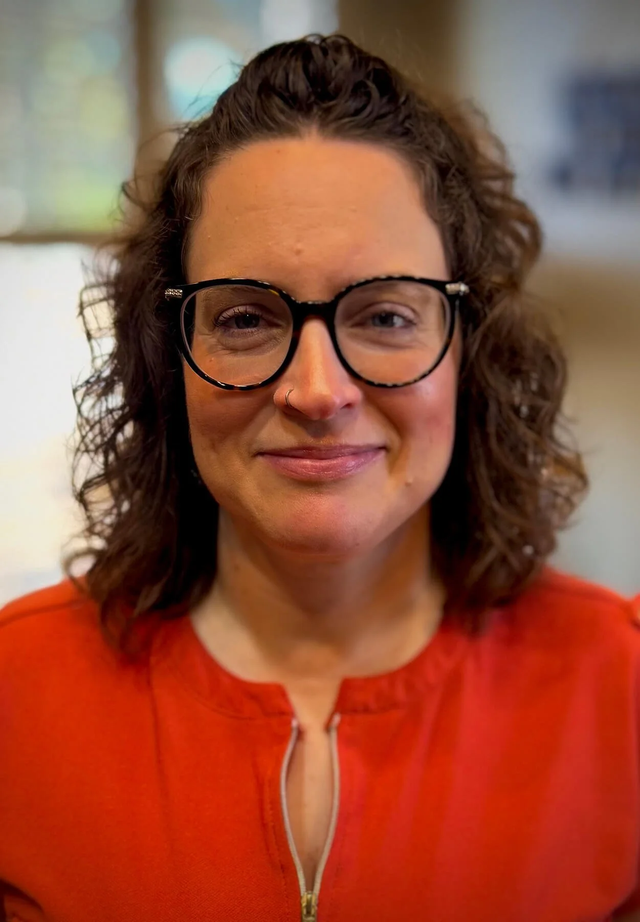 Close-up of a cunselor woman with curly brown hair, wearing black glasses, a nose ring, and a bright orange zip-up top, smiling softly.