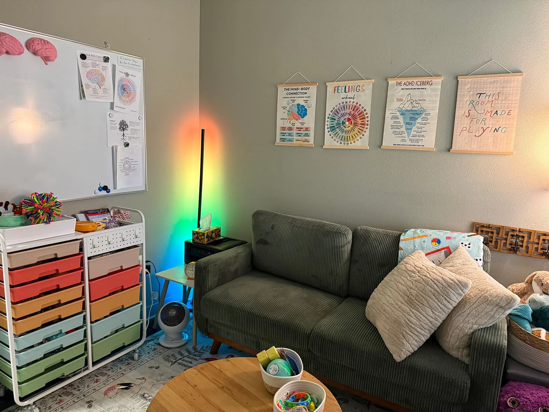 A cozy living room with a green striped sofa, a white cabinet filled with colorful storage drawers, and a whiteboard with various posters on the wall. A small table with bowls of craft supplies and a rainbow-colored light display creates a vibrant atmosphere. There are decorative pillows on the sofa, and the room has a rug with a floral design.