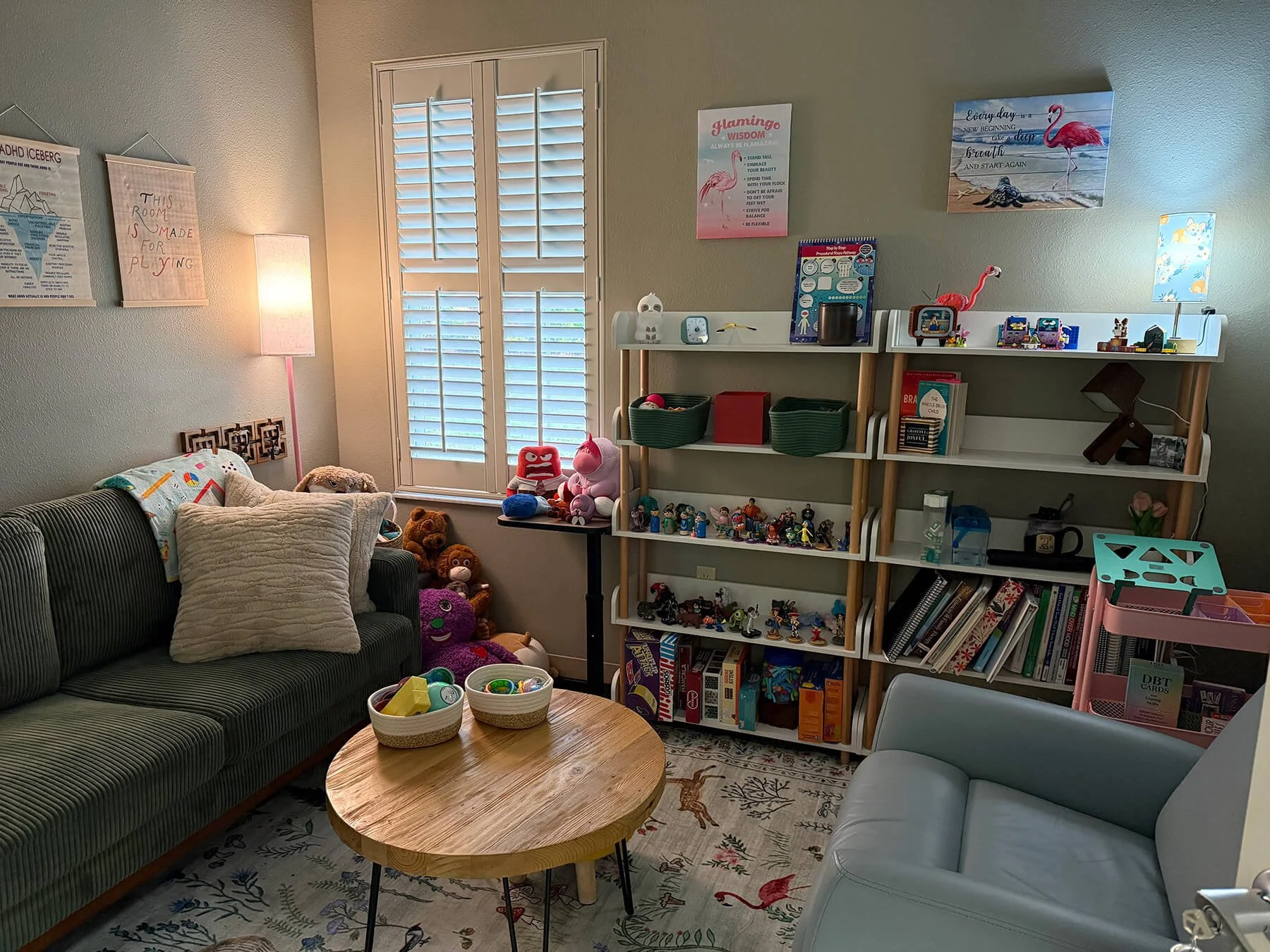 A cozy living room with a dark upholstered sofa topped with pillows, a wooden coffee table with baskets of toys, a white armchair, and a tall shelving unit filled with books, toys, and decorations. The wall has art and posters, and there are window shutters letting in natural light.
