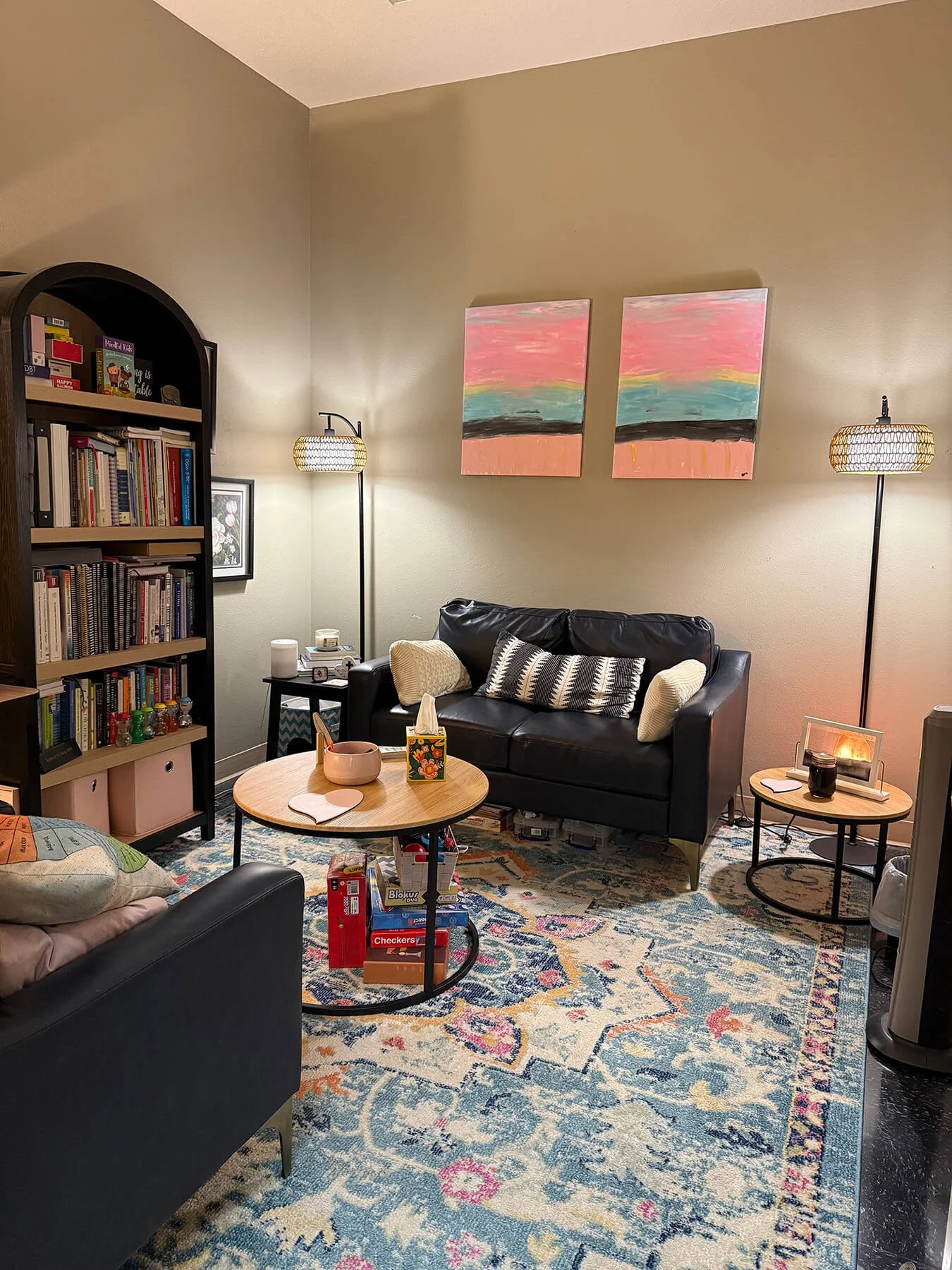 Counselng office with two black sofas, a round wooden coffee table, two lamps, a colorful rug, a bookshelf, and wall art.