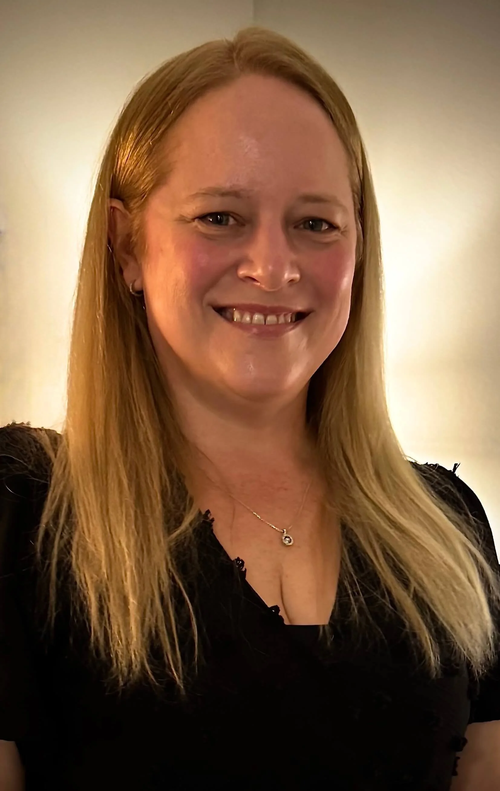 Close-up of a smiling counselor woman with long blonde hair, wearing a black top and a necklace, standing indoors with a plain beige wall in the background.