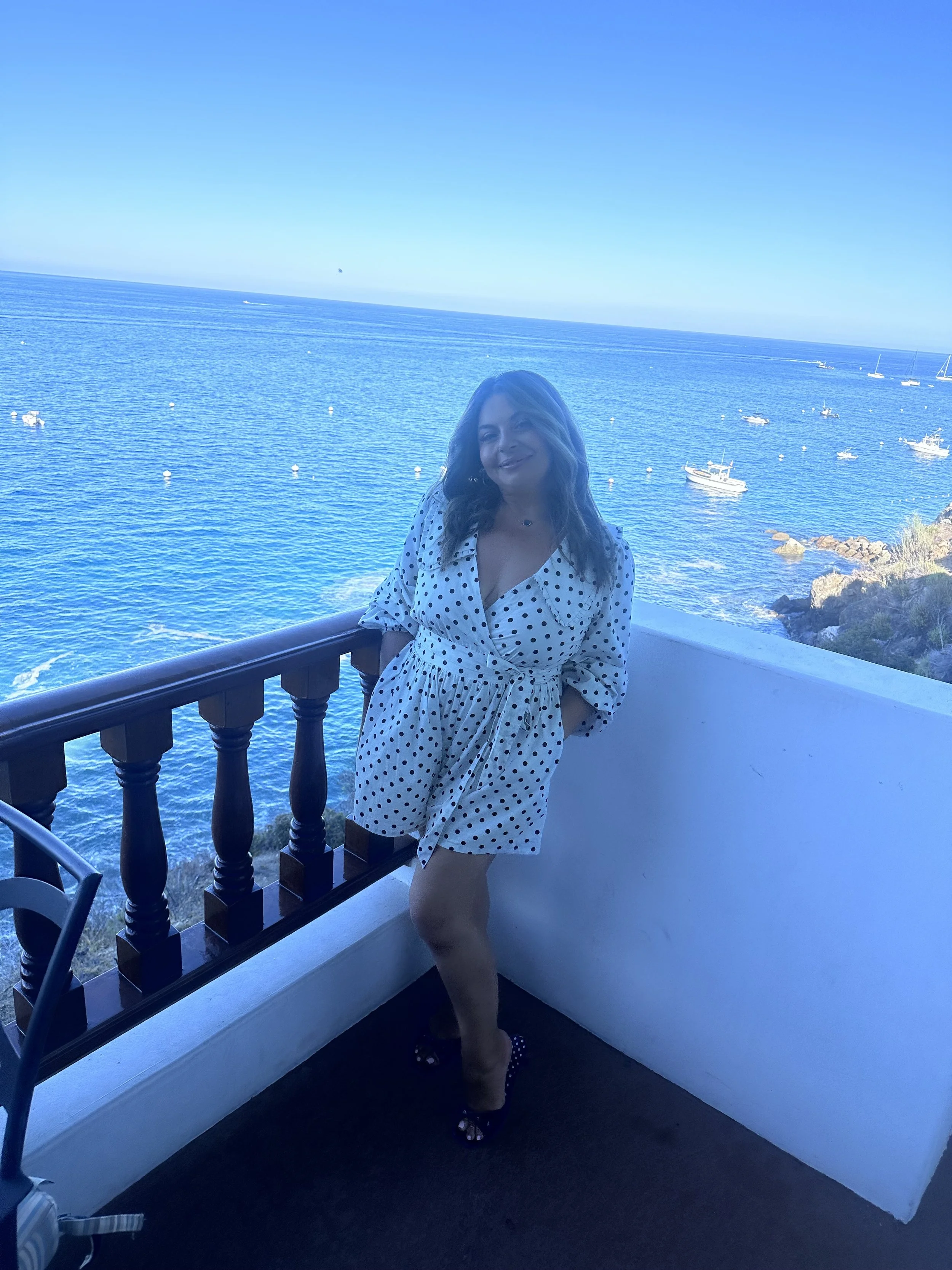 Katherine Karapetian in a white polka dot dress standing on a balcony overlooking the ocean, reflecting calm, freedom, and personal growth. Find Your Sparkk, personal growth.
