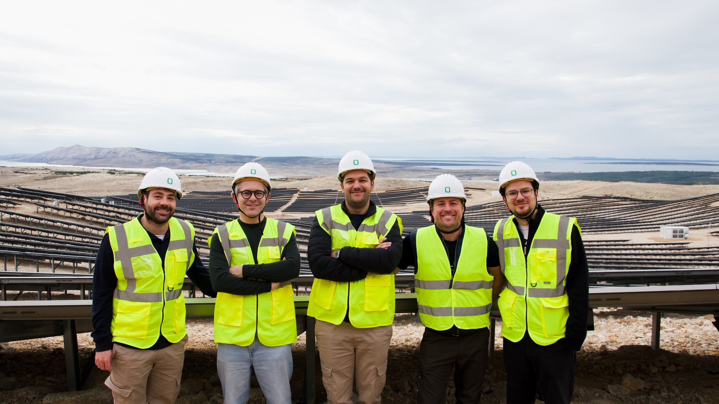 Five construction workers wearing yellow safety vests and white hard hats standing in front of solar panel installation, with a mountain and lake in the background.
