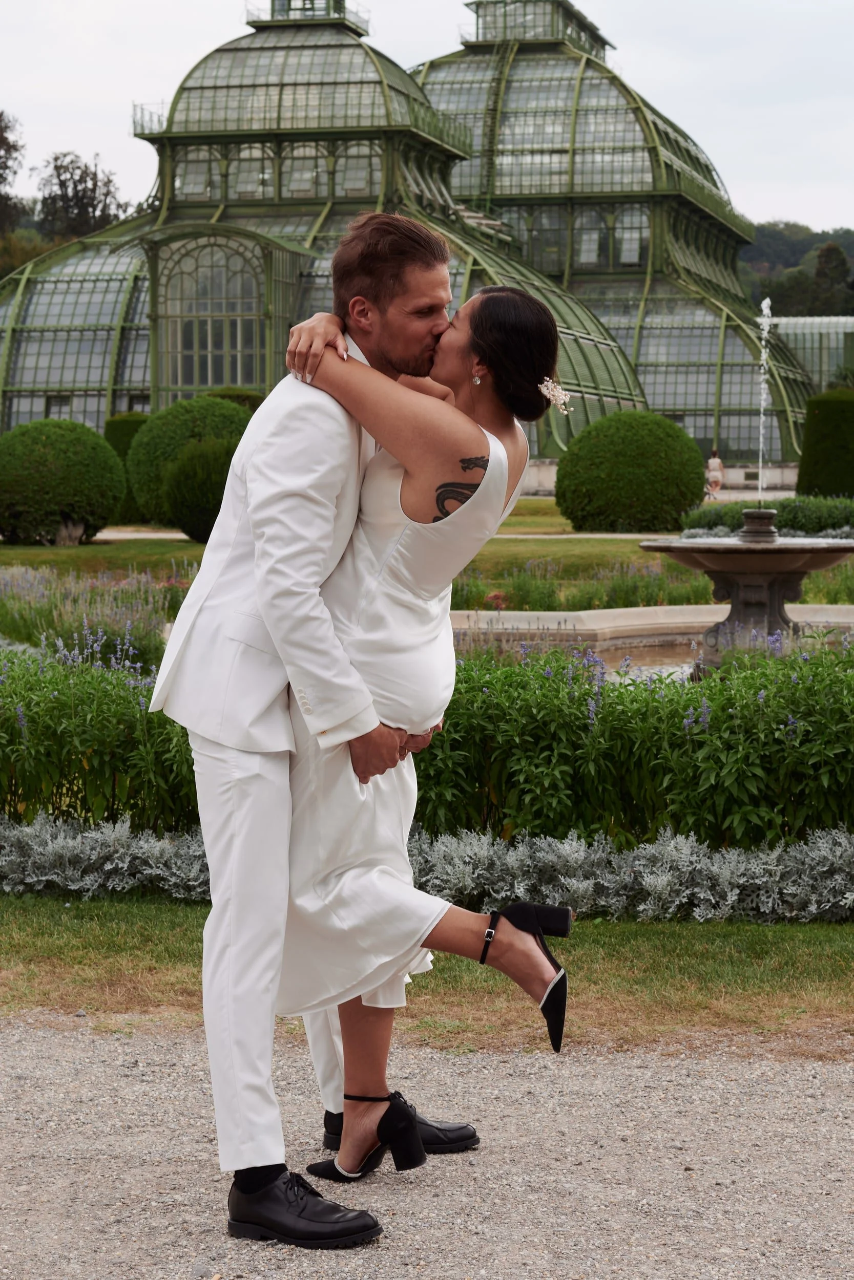 A couple dressed in wedding attire embracing and kissing in front of a greenhouse. The man is wearing a white suit and the woman a white dress with a tattoo on her arm. The woman is lifting her leg and wearing black high heels.