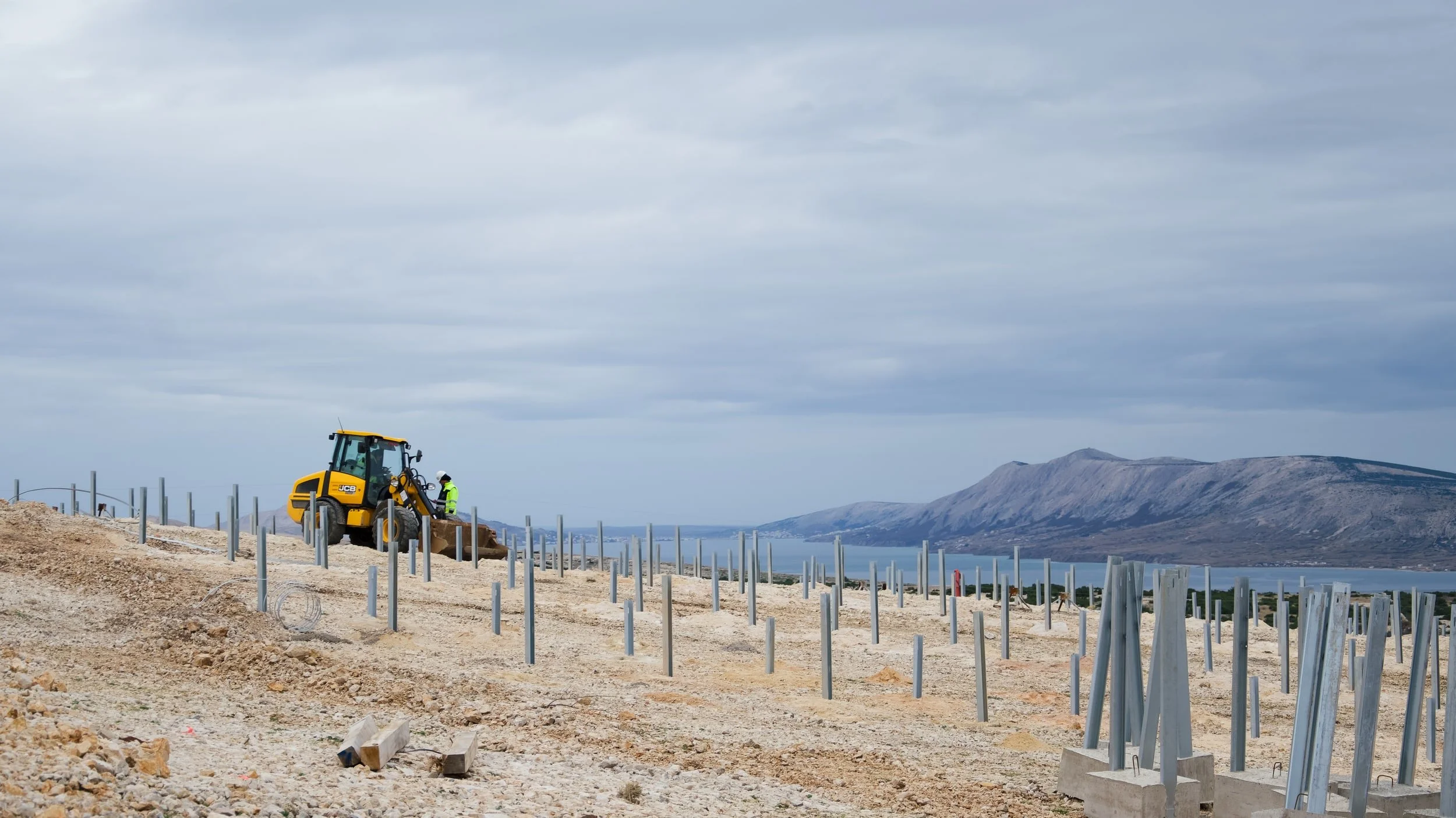 Construction workers and a small JCB excavator working on a hillside construction site with metal fencing posts, overlooking a body of water and mountain range in the background under a cloudy sky.