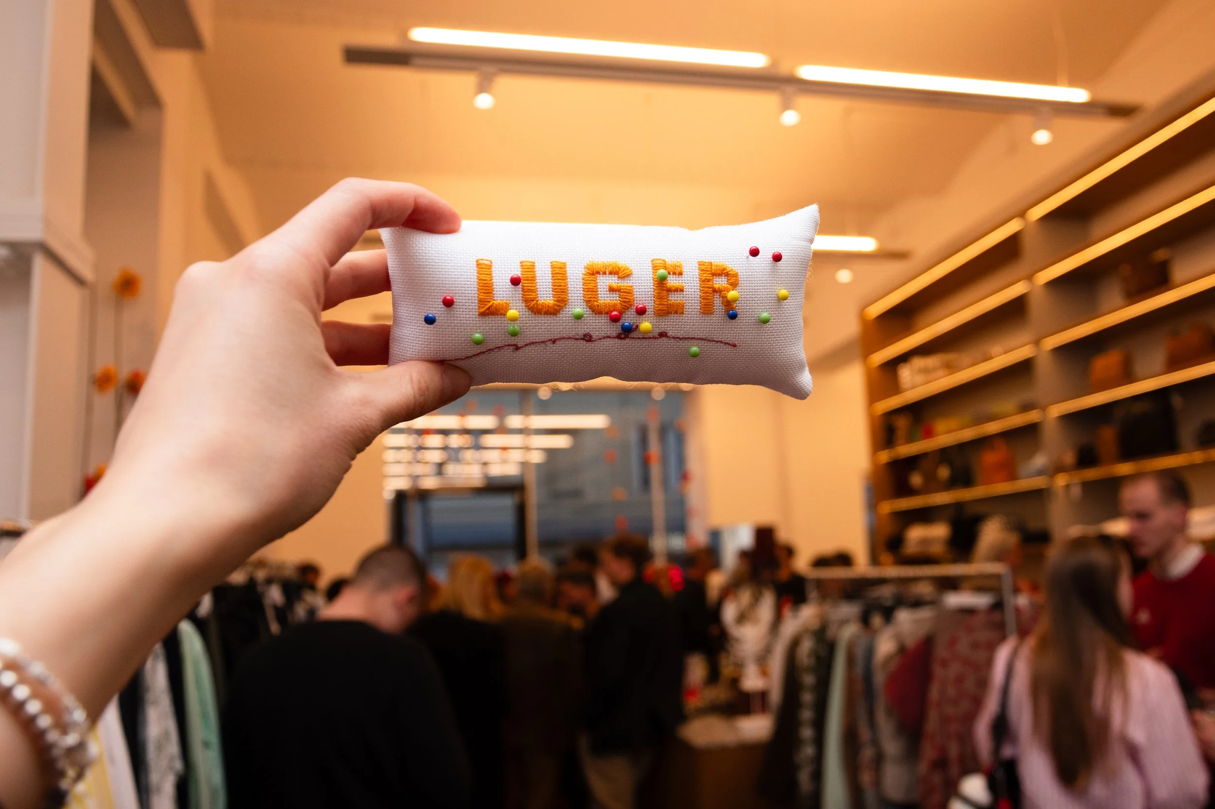 Close-up of a hand holding a small white fabric pillow with the name 'LUGER' embroidered in orange and decorated with multicolored pins, against a store background with shelves and people.