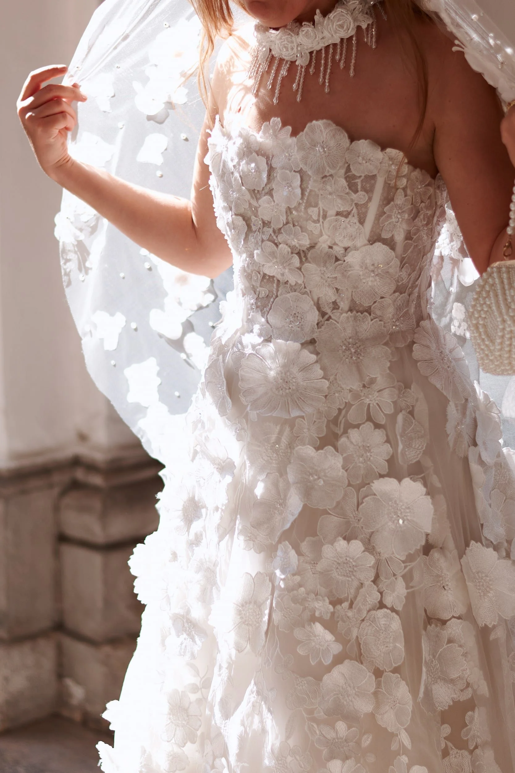 A woman in a wedding dress with intricate floral lace and bead details, holding a sheer, flower-adorned veil. Soft lighting highlights the delicate fabric and embroidery, with part of a stone wall visible in the background.
