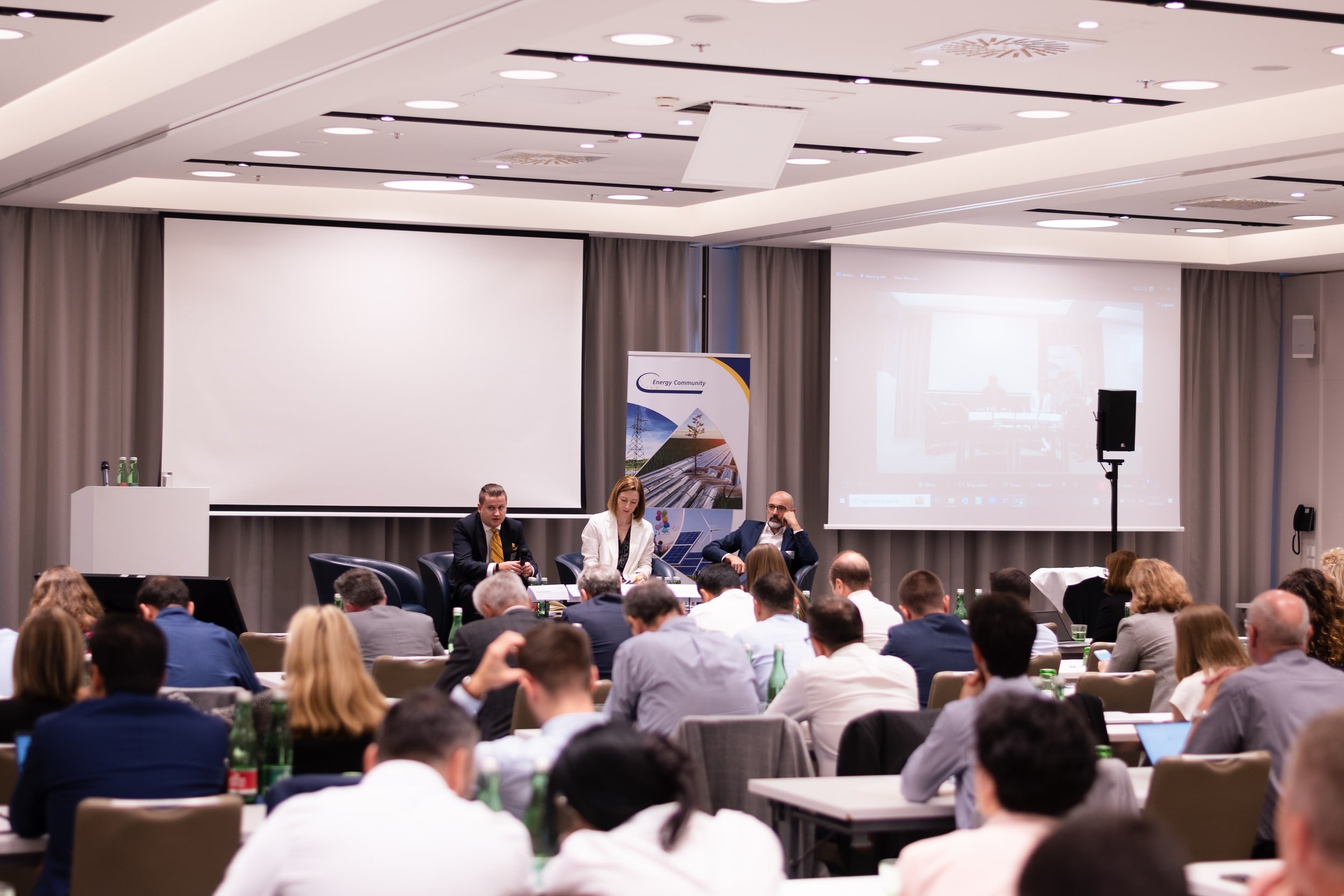 A conference room filled with people seated at tables, listening to a panel of speakers at the front. The room has large presentation screens and curtains on the windows.