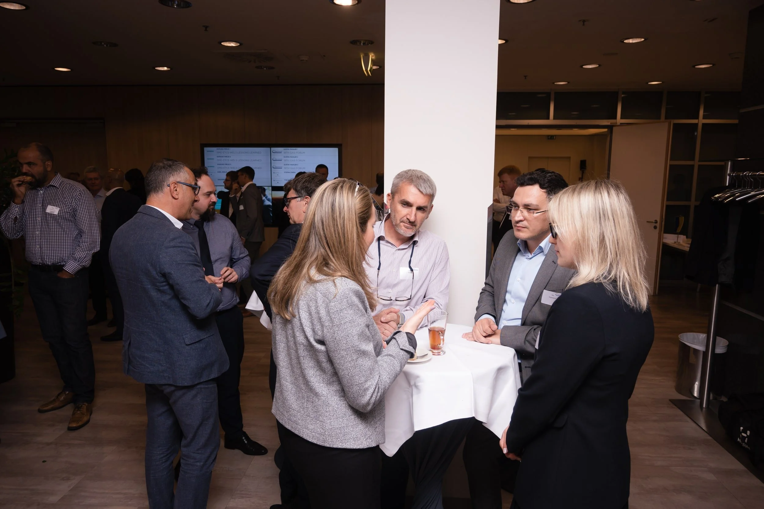A group of people socializing at a networking event or conference in a modern indoor space. Some are engaged in conversation, holding drinks, and wearing business attire.