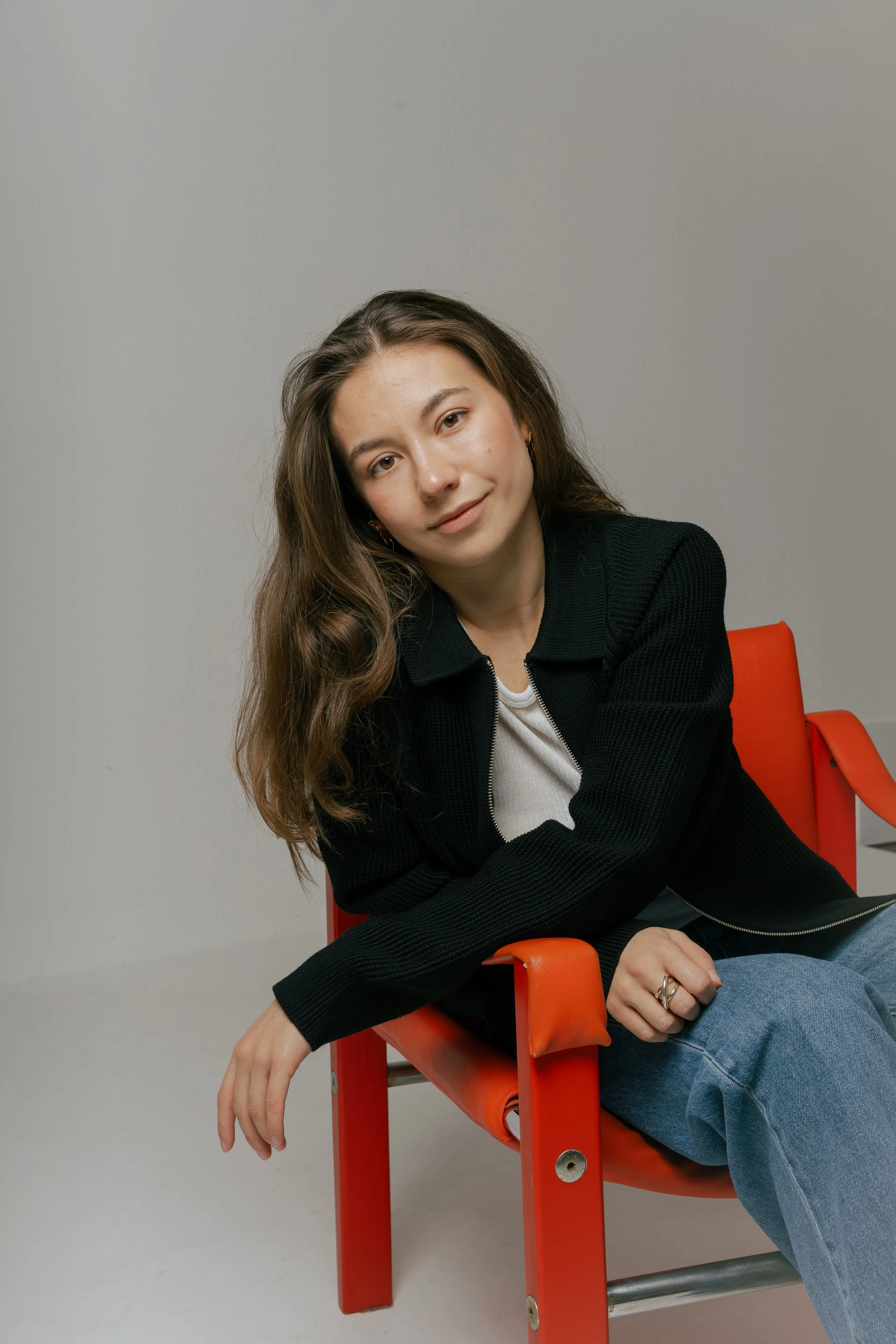 Young woman with long wavy brown hair sitting on an orange chair with a gray background. She wears a black zip-up jacket, a white top, and blue jeans. She is smiling slightly and leaning with her left arm on the armrest of the chair.
