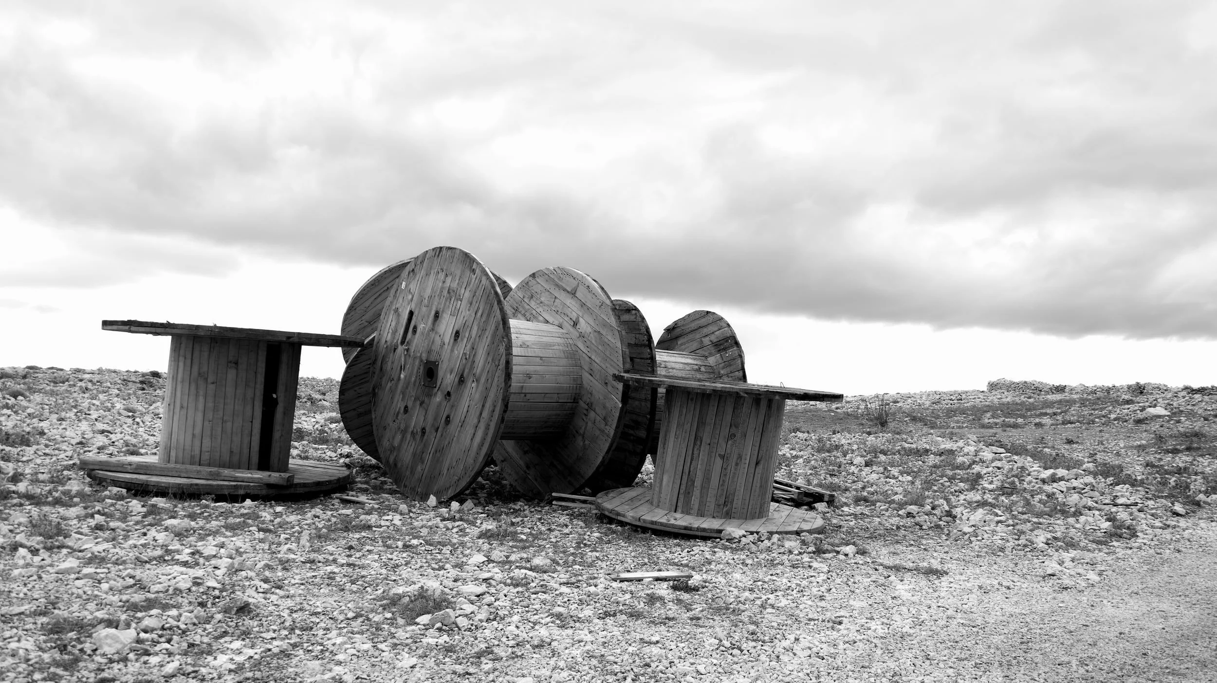 Black and white photo of large wooden spools and benches outdoors on rocky ground under cloudy sky.