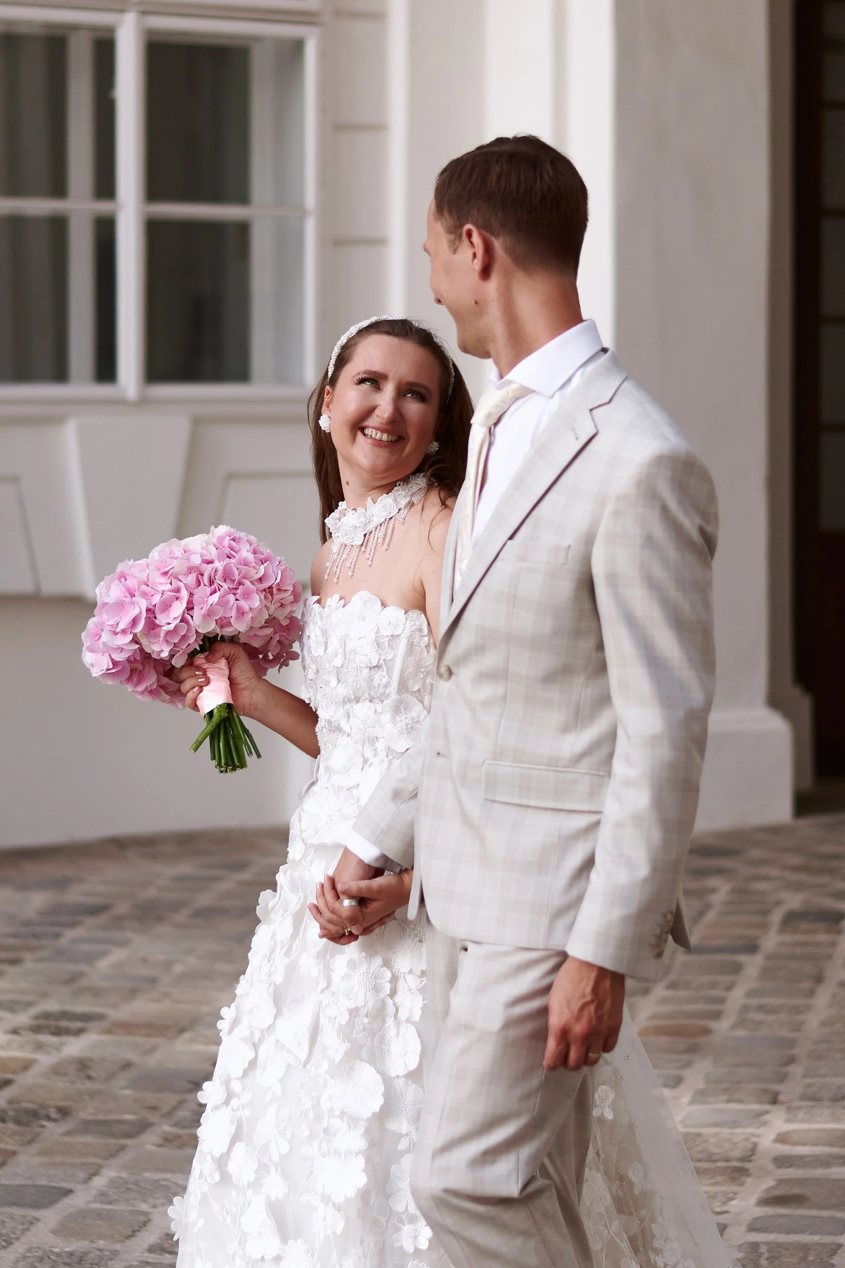 A bride and groom holding hands and smiling at each other indoors, with the bride holding a pink hydrangea bouquet, wearing wedding dresses and suits.