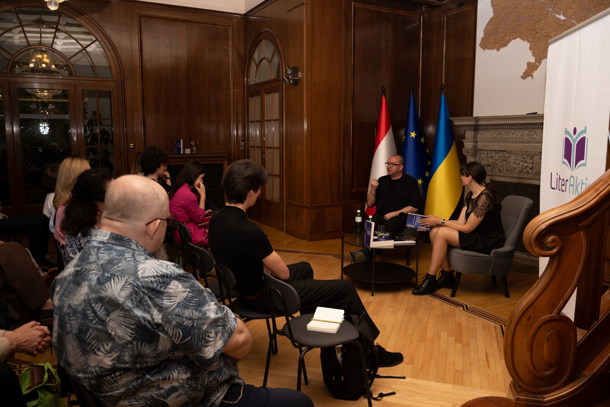 A panel discussion taking place in a wood-paneled room with multiple people sitting and listening. Behind the panel, there are flags of Austria, the European Union, and Ukraine, along with a partially visible banner with a logo and text. The room fea
