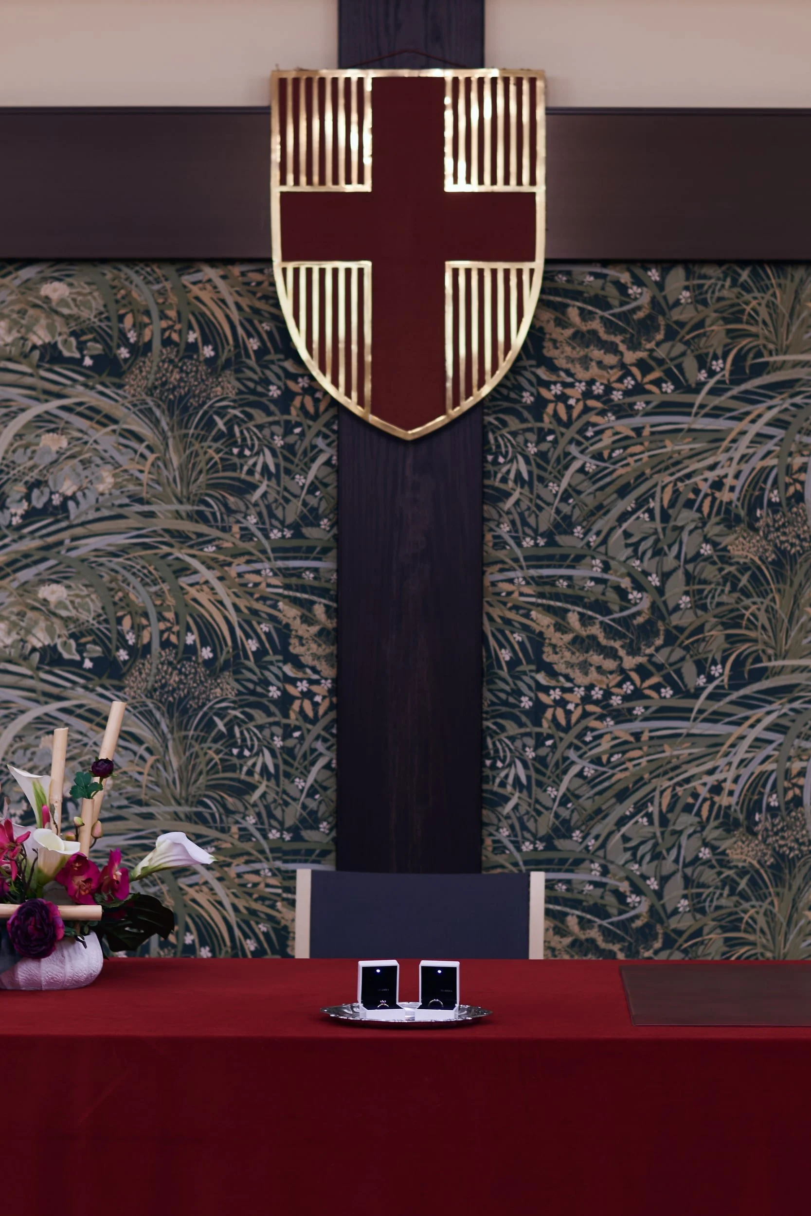 A room decorated with a large crest featuring a red cross on a shield, mounted on a dark wooden panel. There is a table covered with a red cloth, holding two open jewelry boxes. To the left, a bouquet of flowers in a cloth wrap. The background has pa