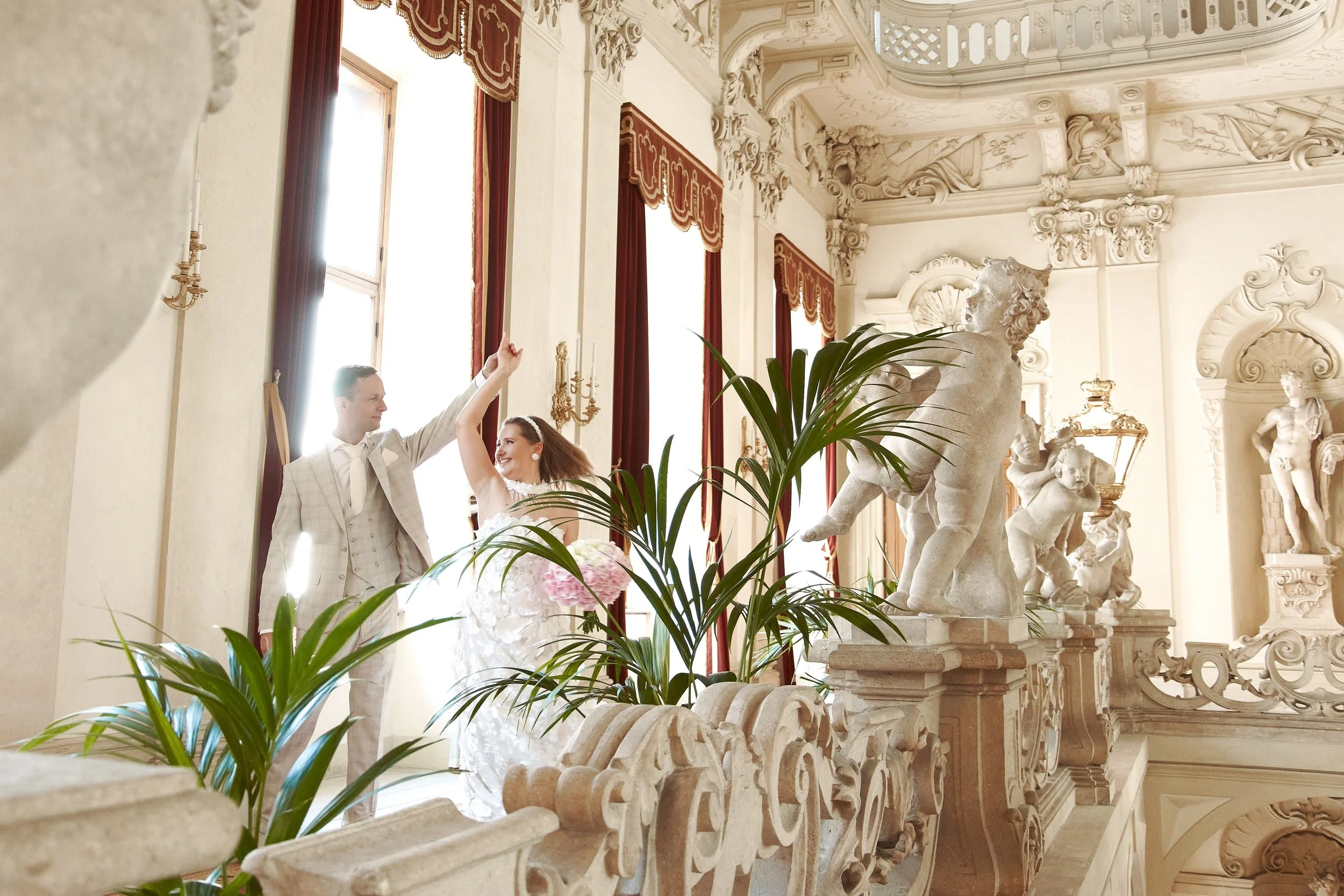 A bride and groom dancing inside a grand, ornate hall with high windows and detailed sculptures of horses and cherubs.