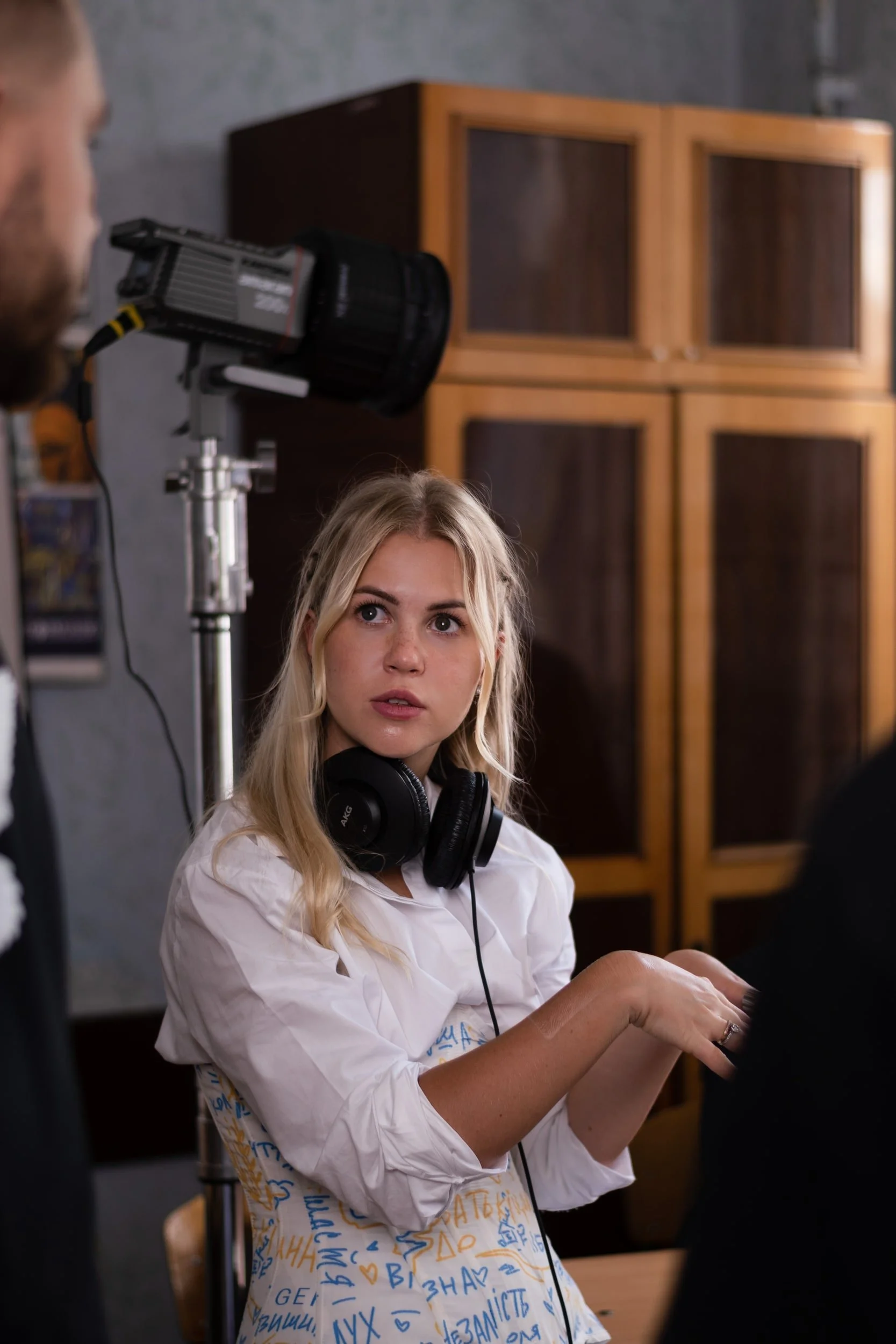 A woman with blonde hair wearing a white shirt with blue and yellow symbols and writing, and large black headphones around her neck, appears to be in a photo shoot or filming setting. She is looking slightly upward and has a focused expression. A per