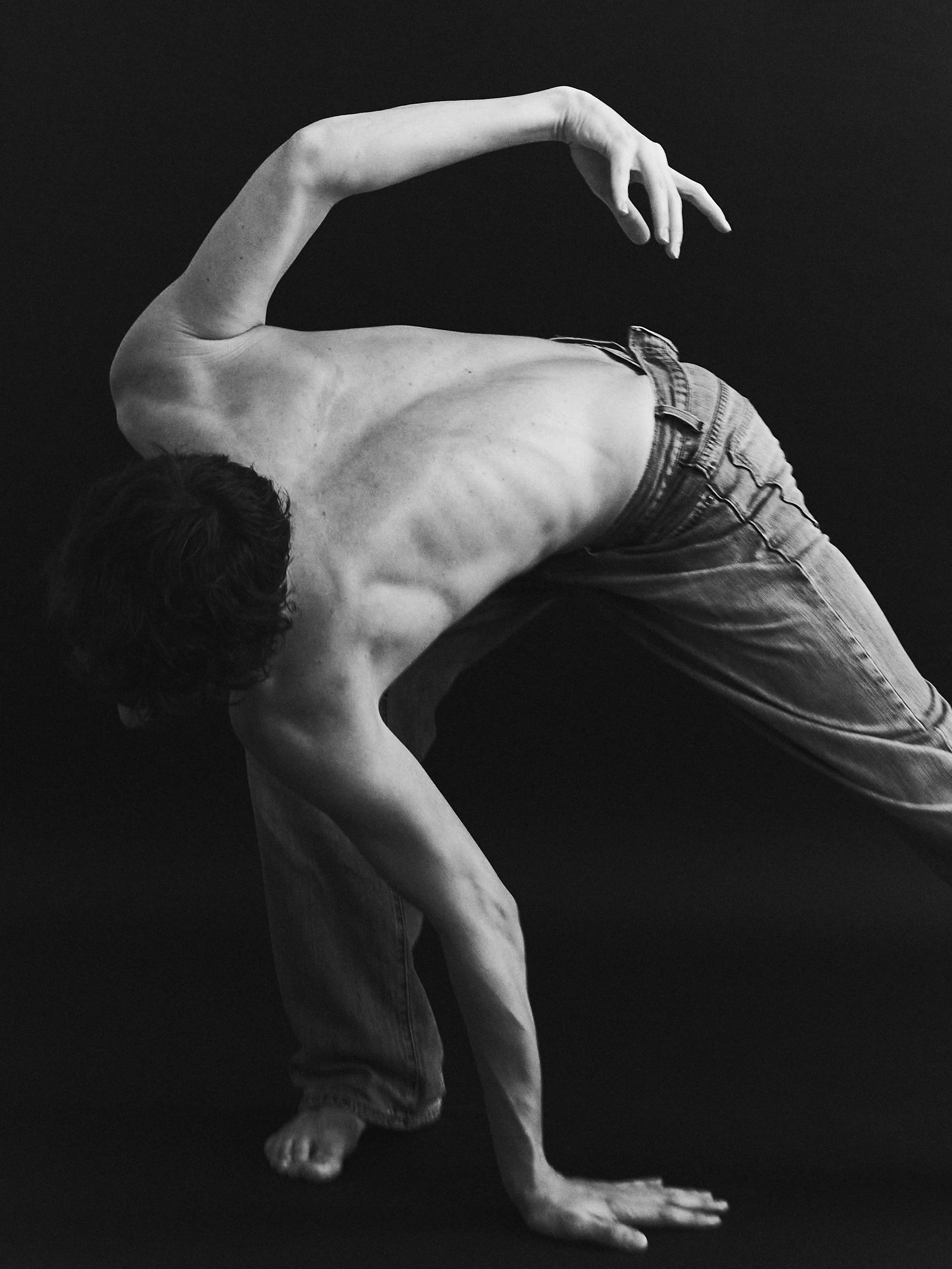 A black and white portrait of a dancer with a bare upper body, wearing jeans, bending over with their head down, one arm reaching forward and the other arm bent, against a dark background.
