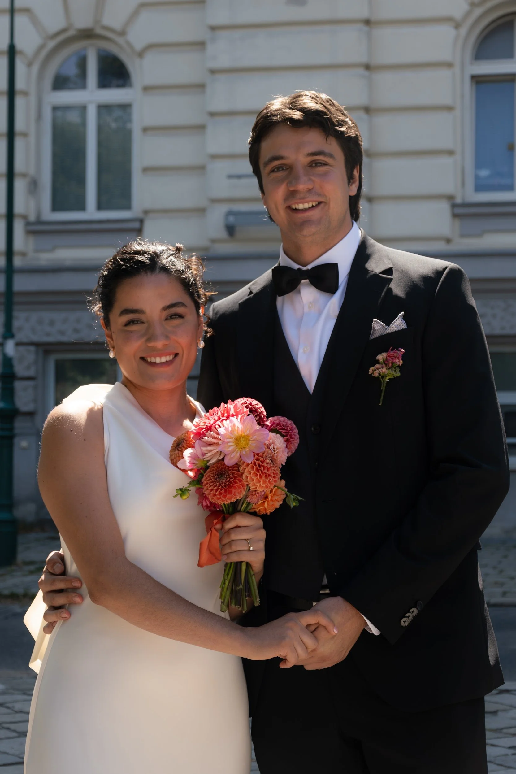 A smiling couple dressed in formal attire, holding a bouquet of pink and orange flowers outside a historic building with large arched windows.