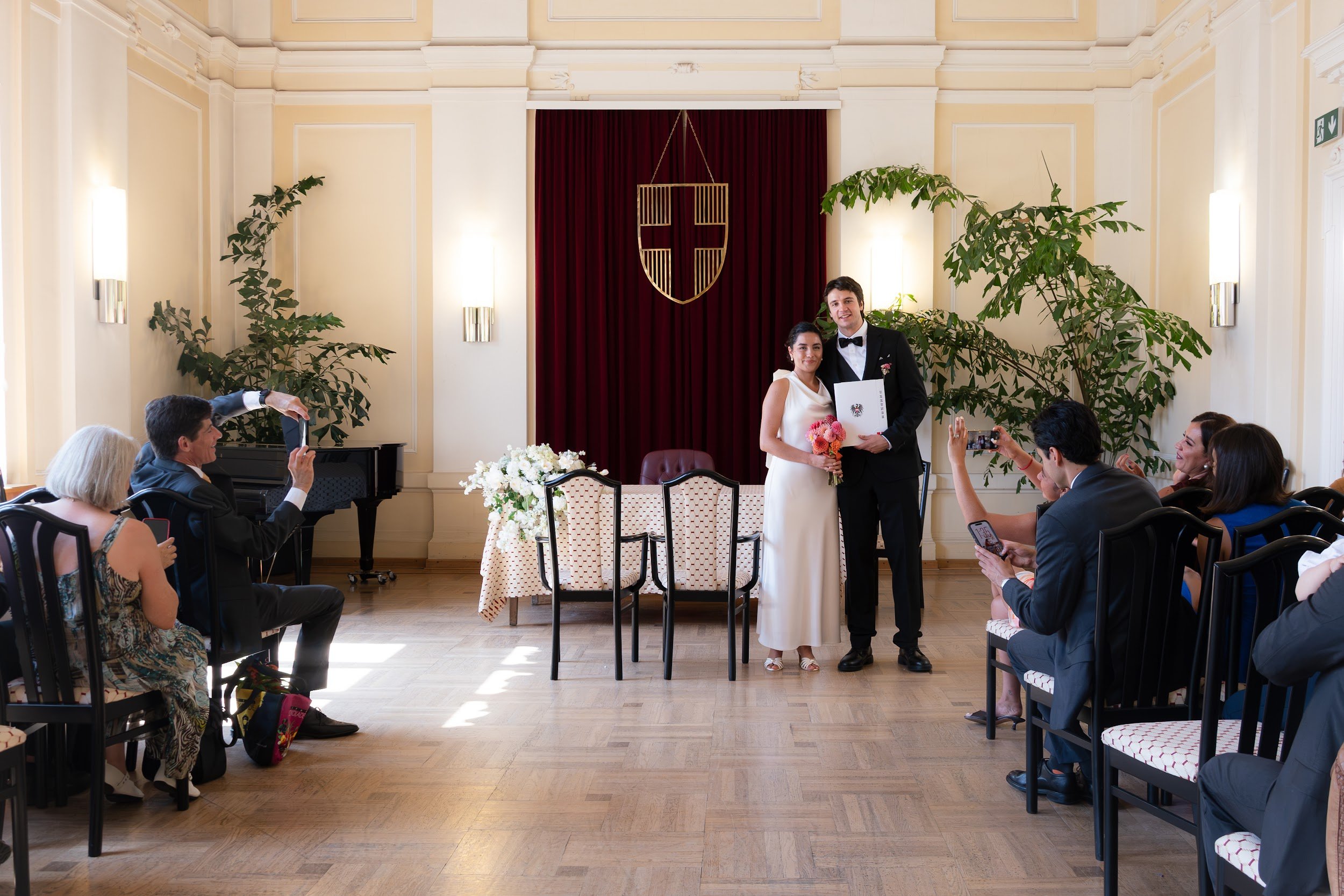 A bride and groom stand together in front of a red curtain with a shield emblem, surrounded by guests taking photos during a wedding ceremony inside a bright, decorated room with cream walls and large potted plants.