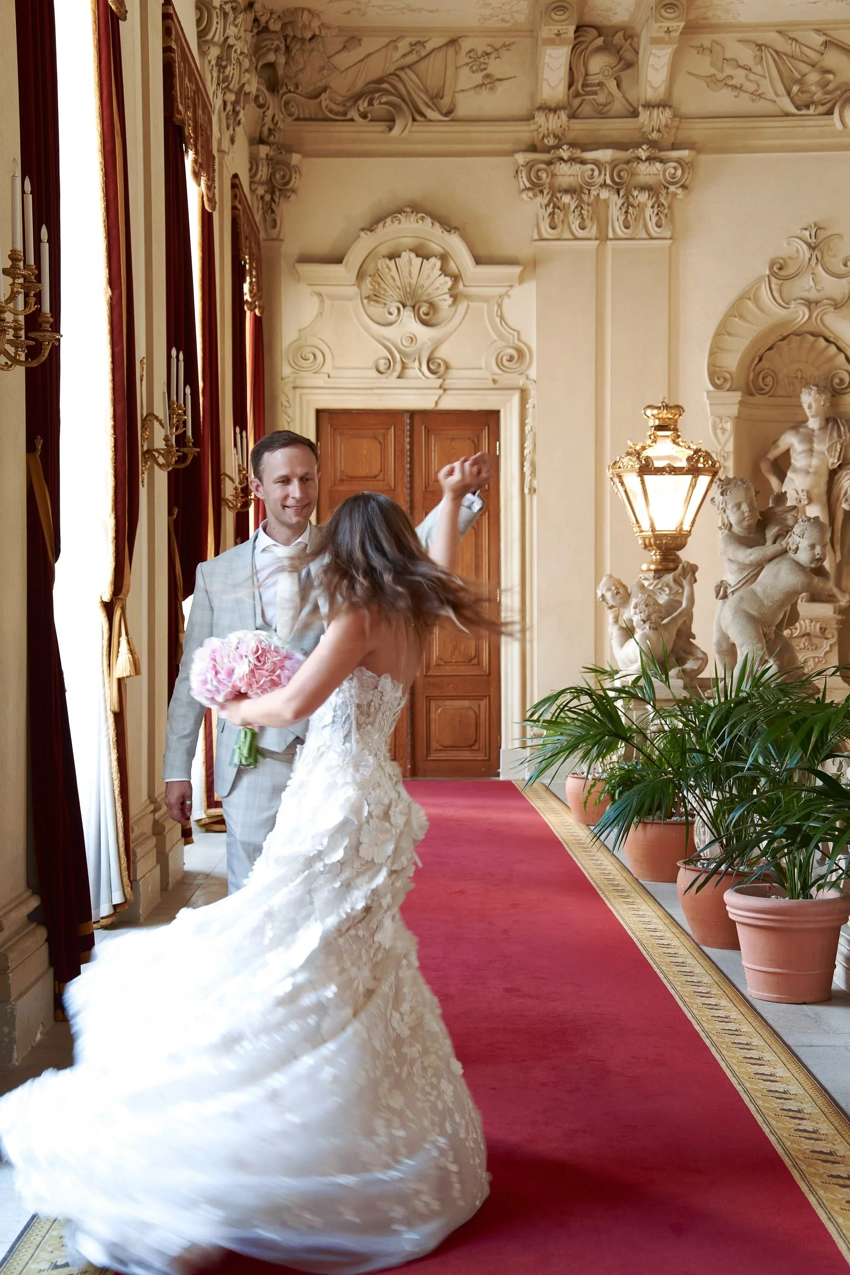 A bride in a white wedding gown dancing with a groom in a light-colored suit in a decorated, elegant room with ornate sculptures, golden wall decor, red curtains, and potted plants.