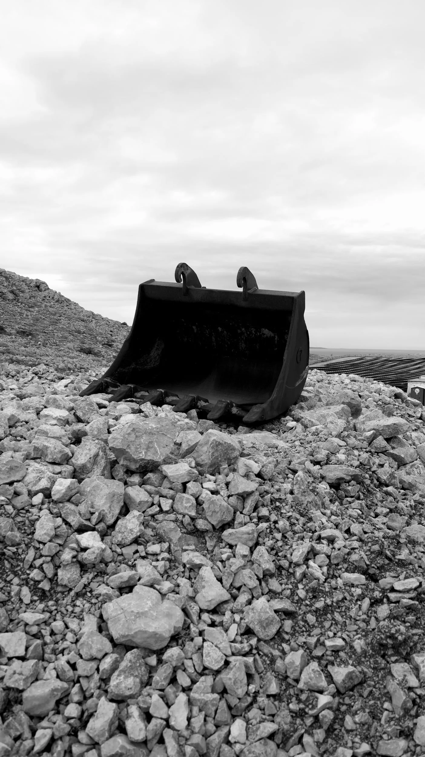 A large excavator bucket lying on rocky terrain under a cloudy sky.
