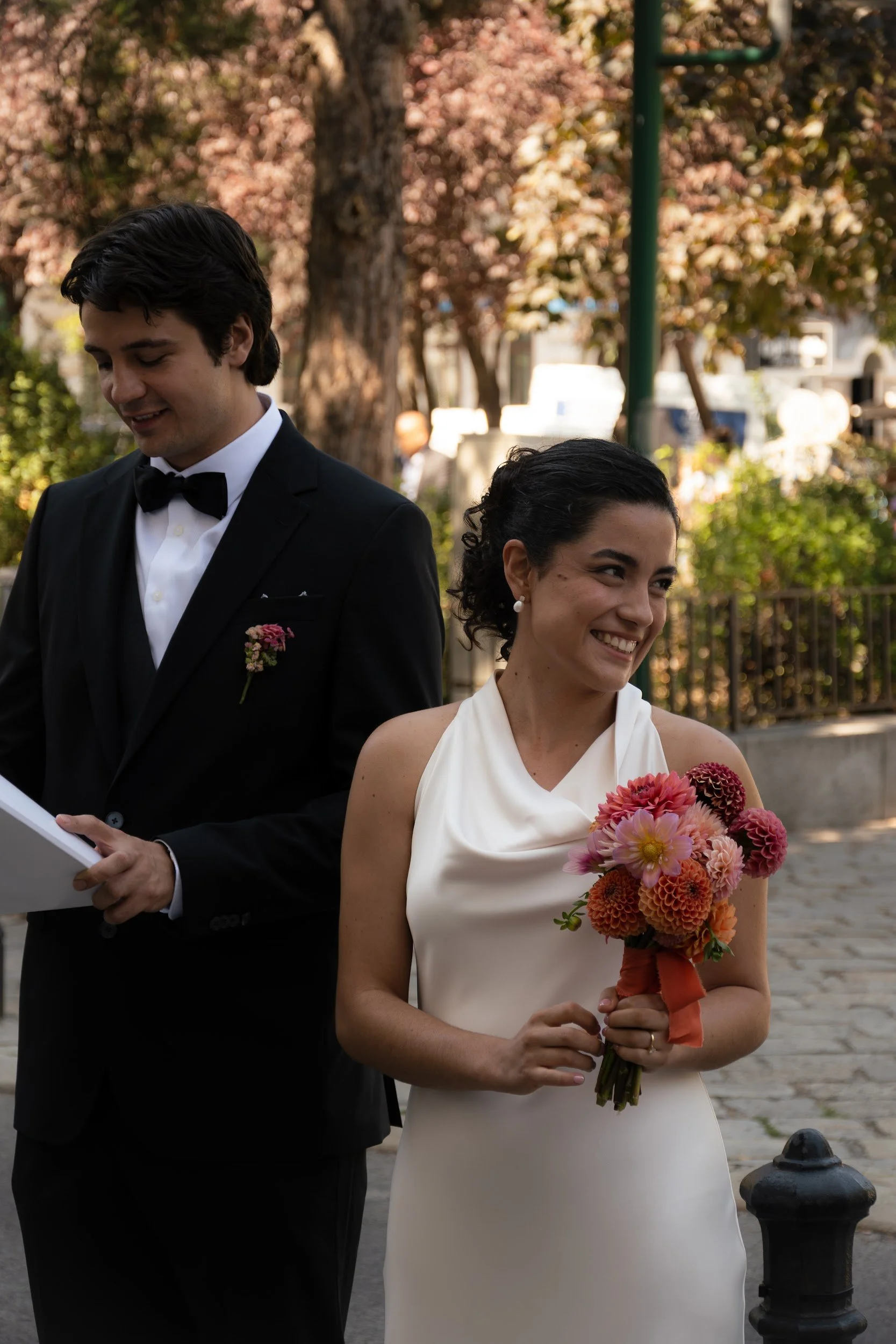 A couple at a wedding or special event: man in a black tuxedo with bow tie, woman in a white dress, holding a bouquet of pink, orange, and red flowers.