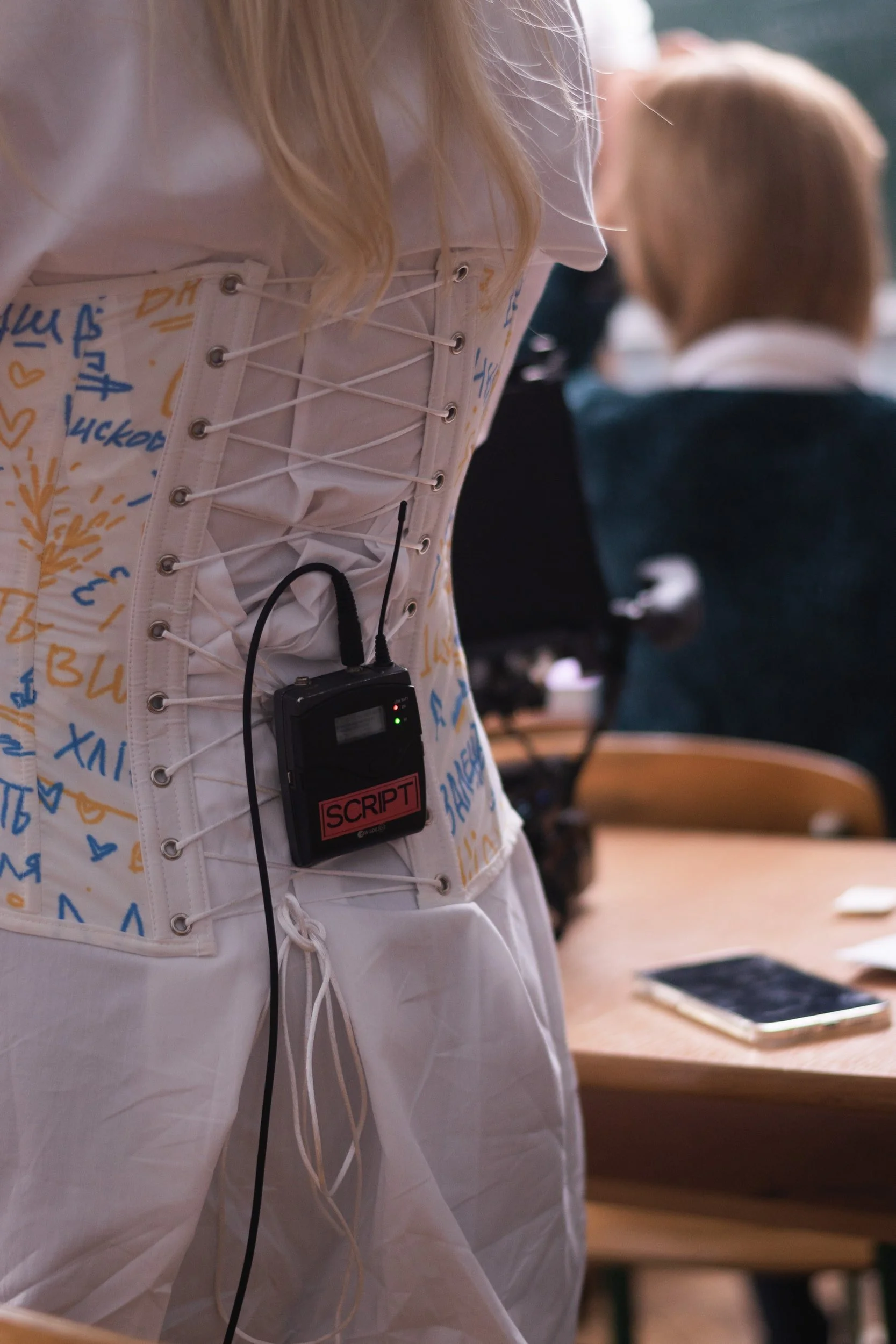 Close-up of a person's torso wearing a corset with laces and metal eyelets, with a small black electronic device labeled 'SCRIPT' attached to the front, in a classroom setting with other students and desks visible in the background.