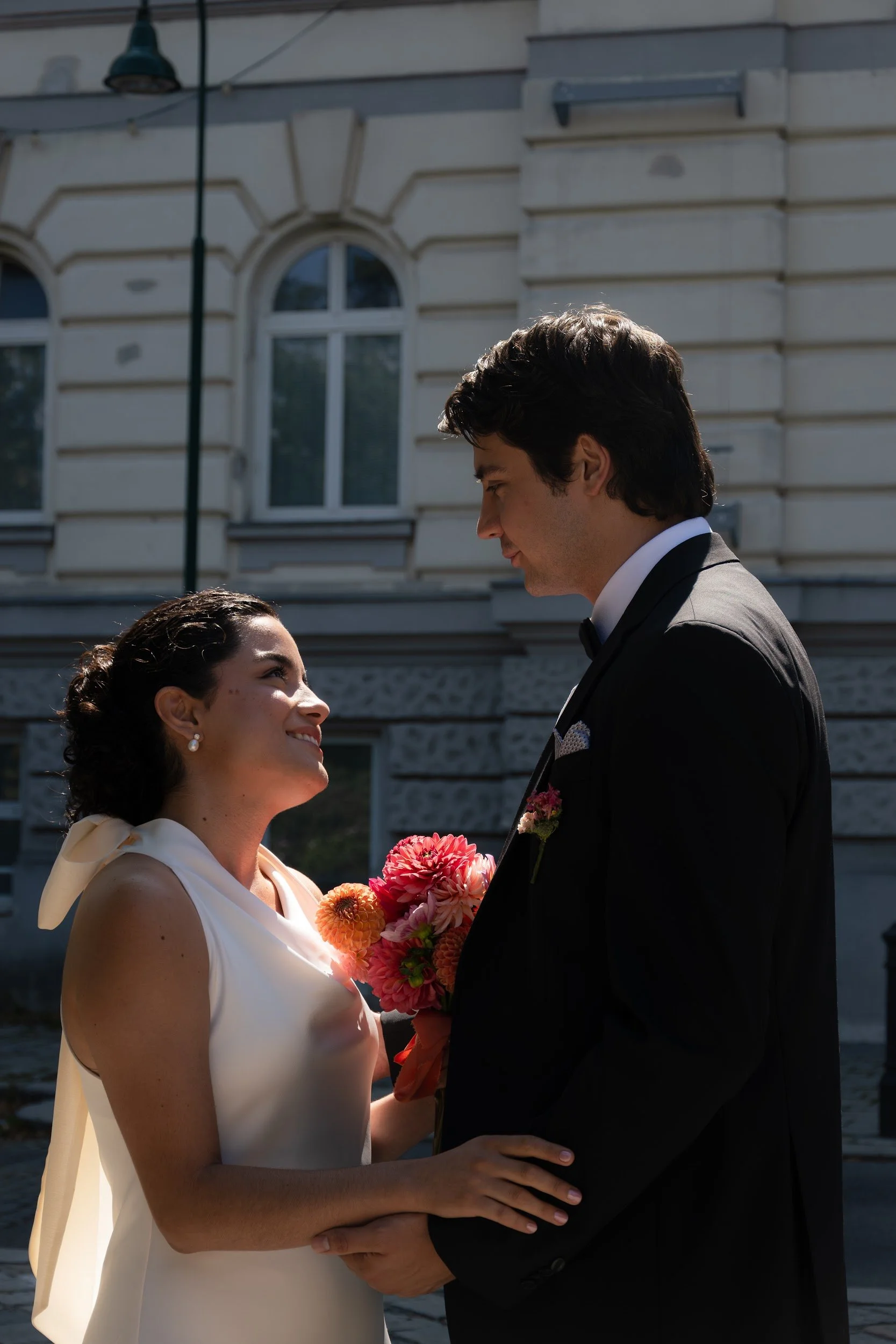 A couple dressed in elegant attire gazes at each other during a wedding ceremony outdoors, with the bride holding a bouquet of colorful flowers and the groom looking at her lovingly.