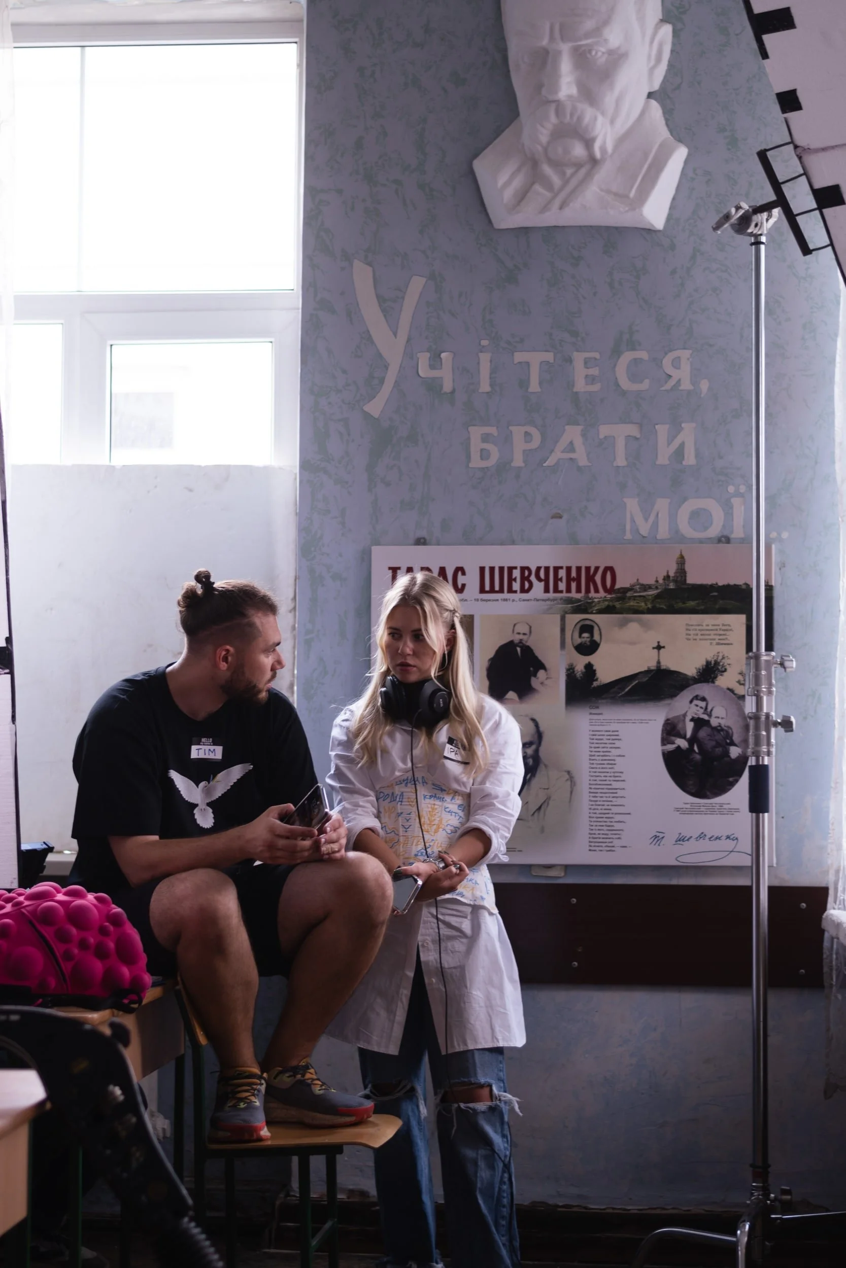Two young people sitting on chairs, one man and one woman, engaged in a conversation, with a woman standing in front of them. The background includes Ukrainian language posters, a portrait of a man, and a window, with a lighting setup nearby.