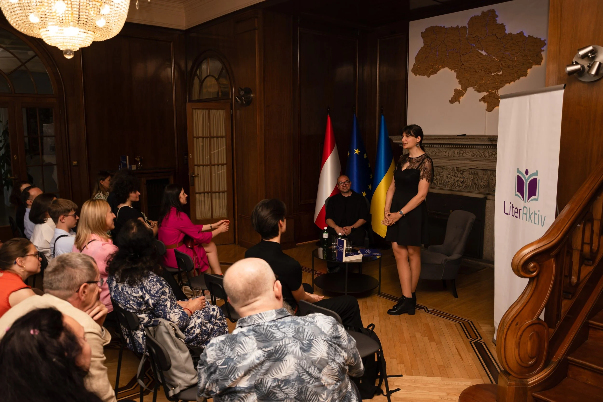 Inside a wood-paneled room with a large map of Ukraine on the wall, a woman stands while speaking to an audience. Flags of Ukraine, the European Union, and Austria are displayed behind her. Audience members are seated, listening attentively.