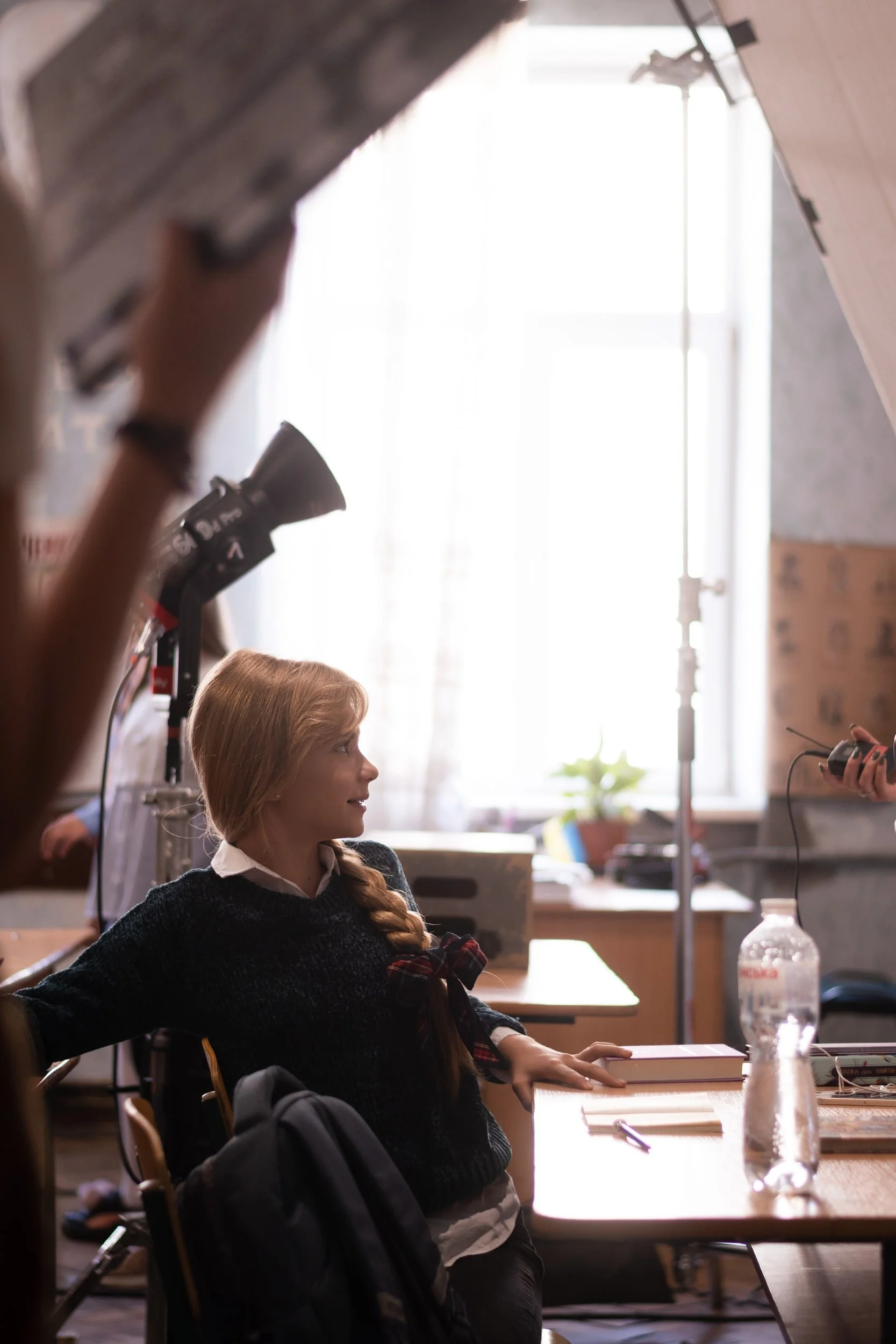 A young girl with long braided hair wearing a dark sweater seated at a desk in a classroom, with her arm resting on the table, a book in front of her, and looking at someone off-camera. In the background, there is a woman holding a tablet and a water