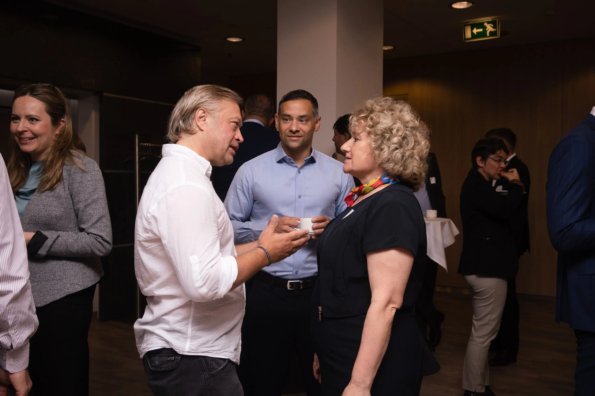 Two men and a woman engaged in conversation at a social event, with other guests in the background.