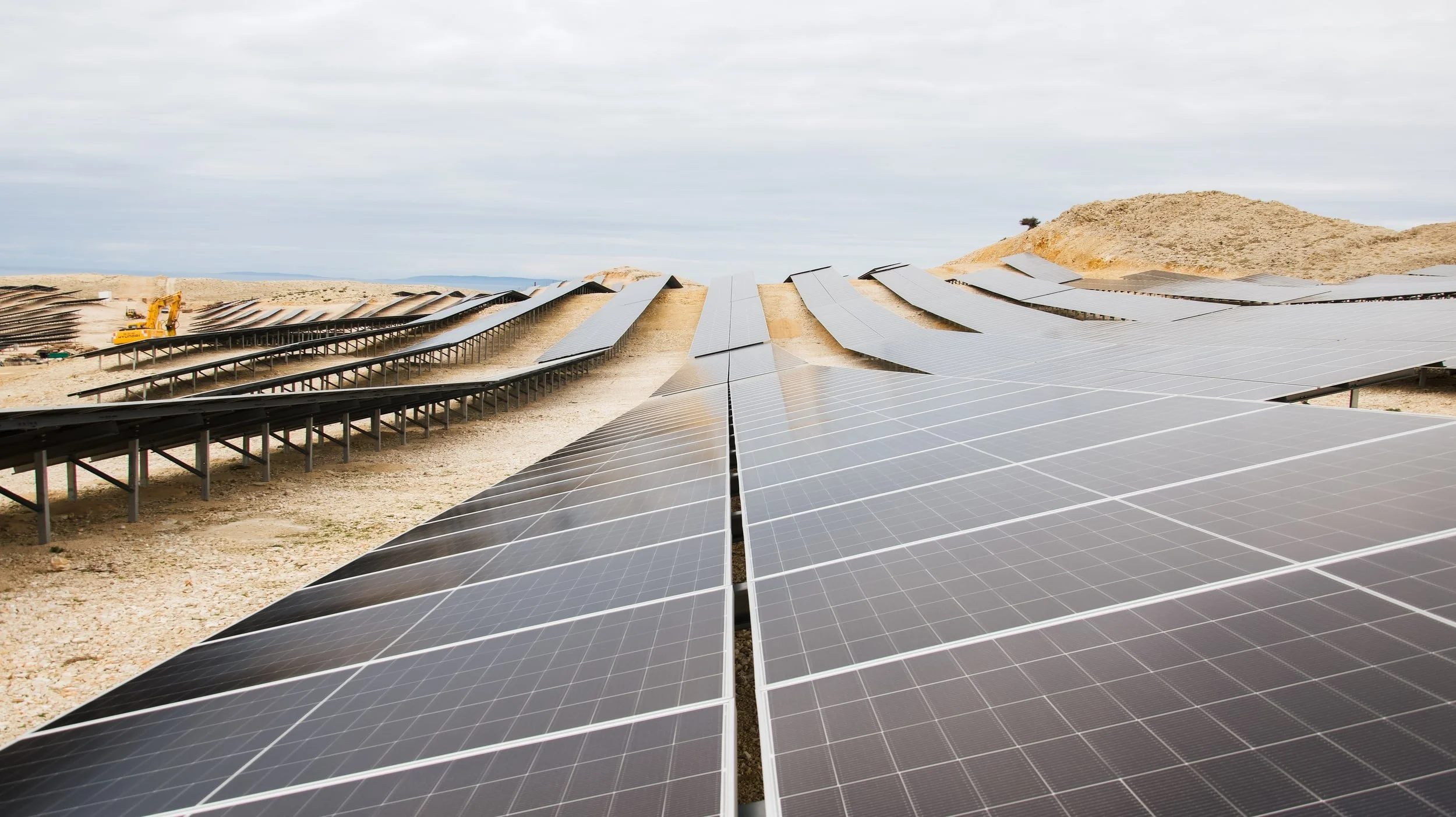 A large solar panel installation on a desert landscape with hills in the background and construction equipment in the distance.