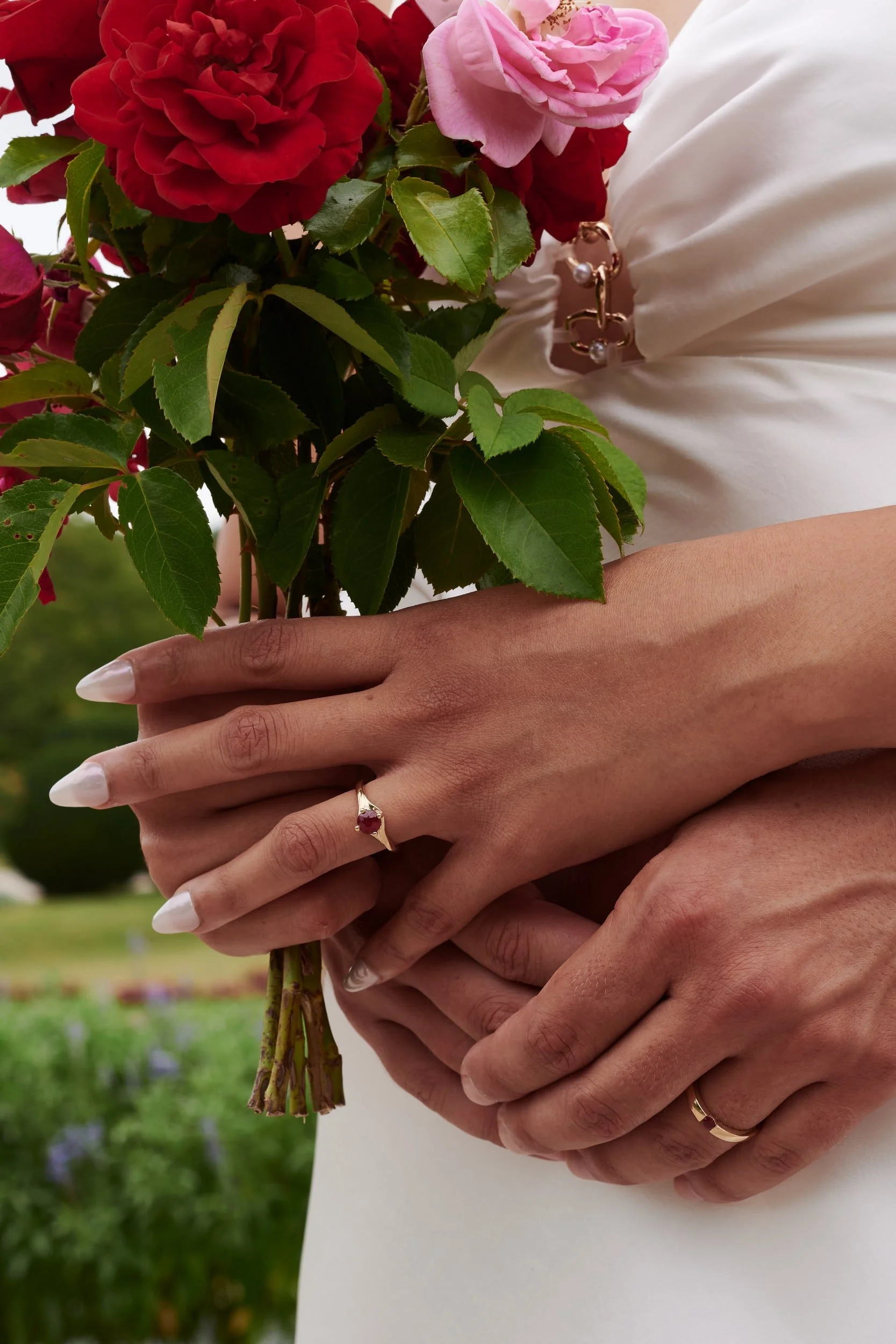 Close-up of two people holding a bouquet of red and pink roses with green leaves, one person wearing a white dress with a gold ring and bracelet, the other person’s hands overlapping, in a park or garden setting.