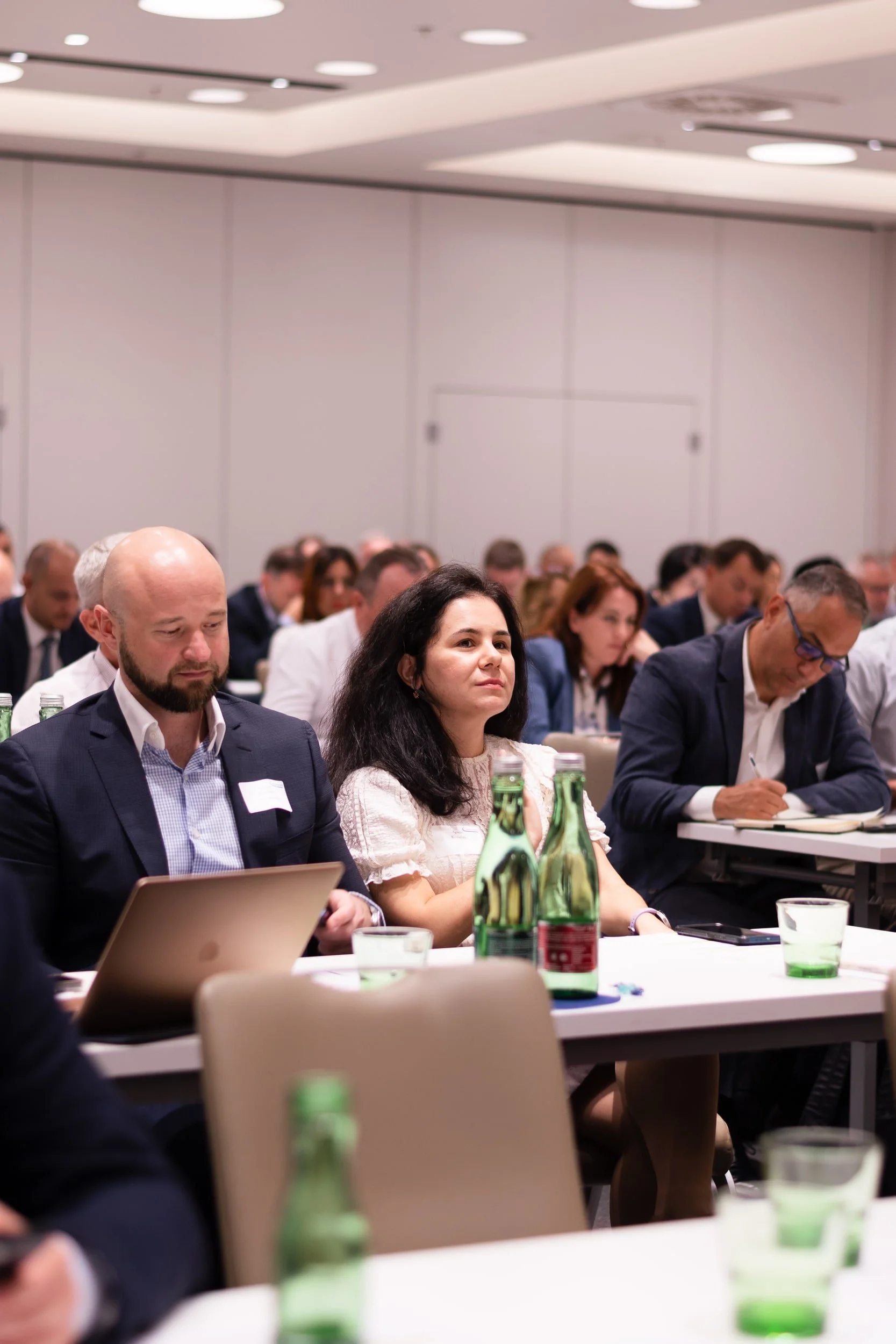 People attending a conference or meeting, sitting at tables with bottles and glasses, some taking notes or using laptops in a large, well-lit room.