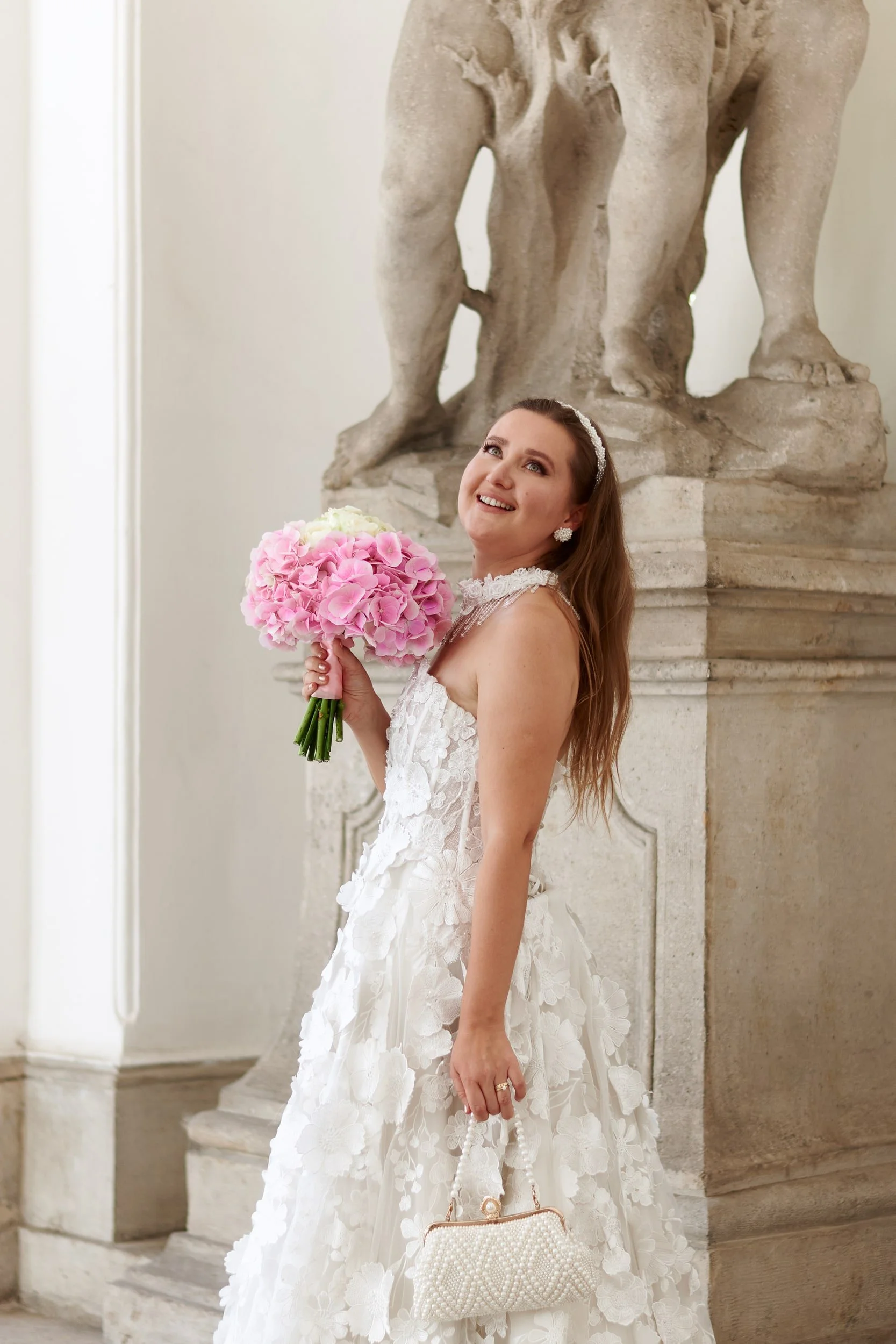 A bride in a white wedding gown holding a bouquet of pink and white roses, standing in front of a large stone statue of a lion.