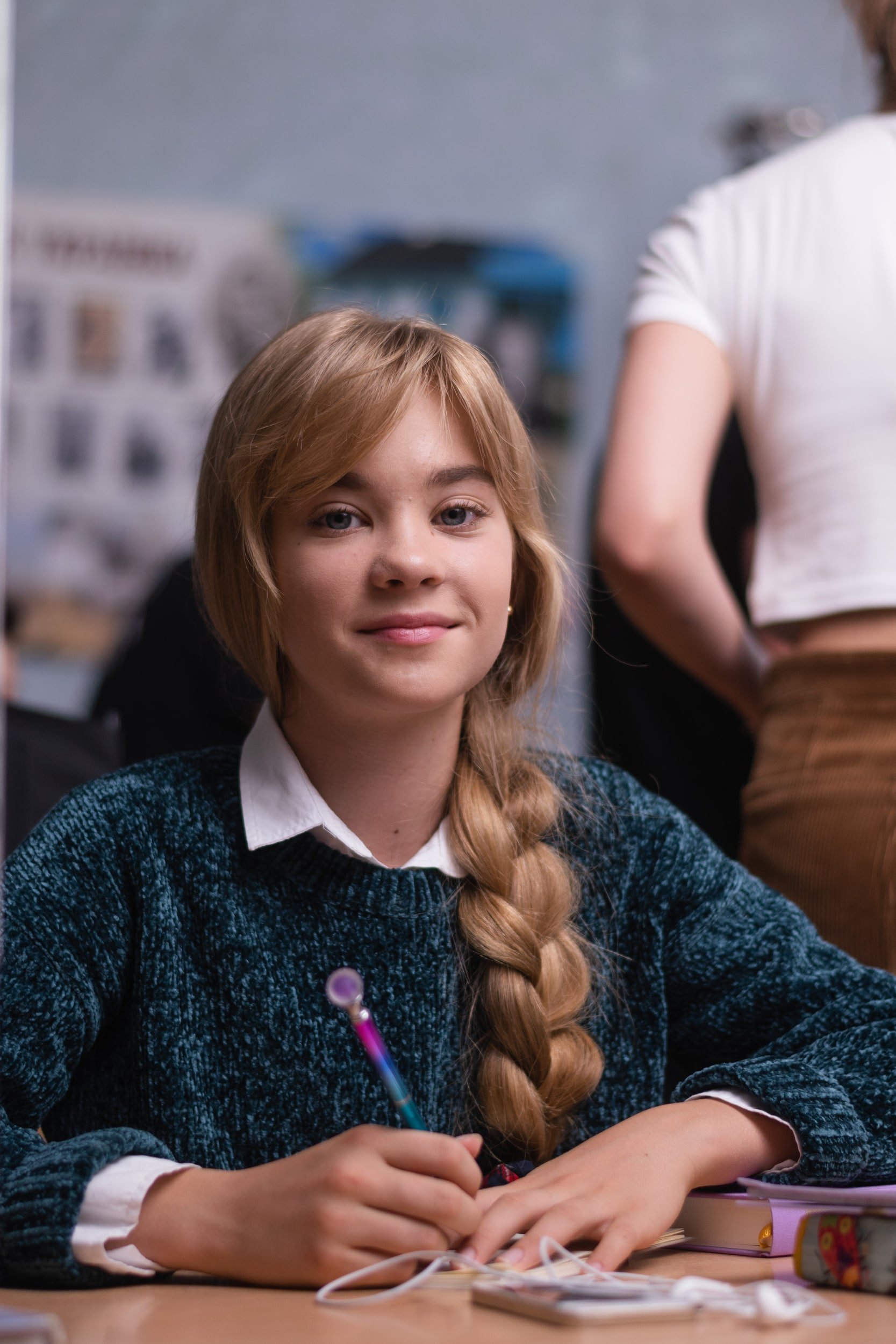 A young woman with long blonde hair styled in a braid, wearing a dark blue textured sweater over a white collared shirt, sits at a desk with a pink notebook, colorful pens, and other stationery items. She looks directly at the camera with a slight sm