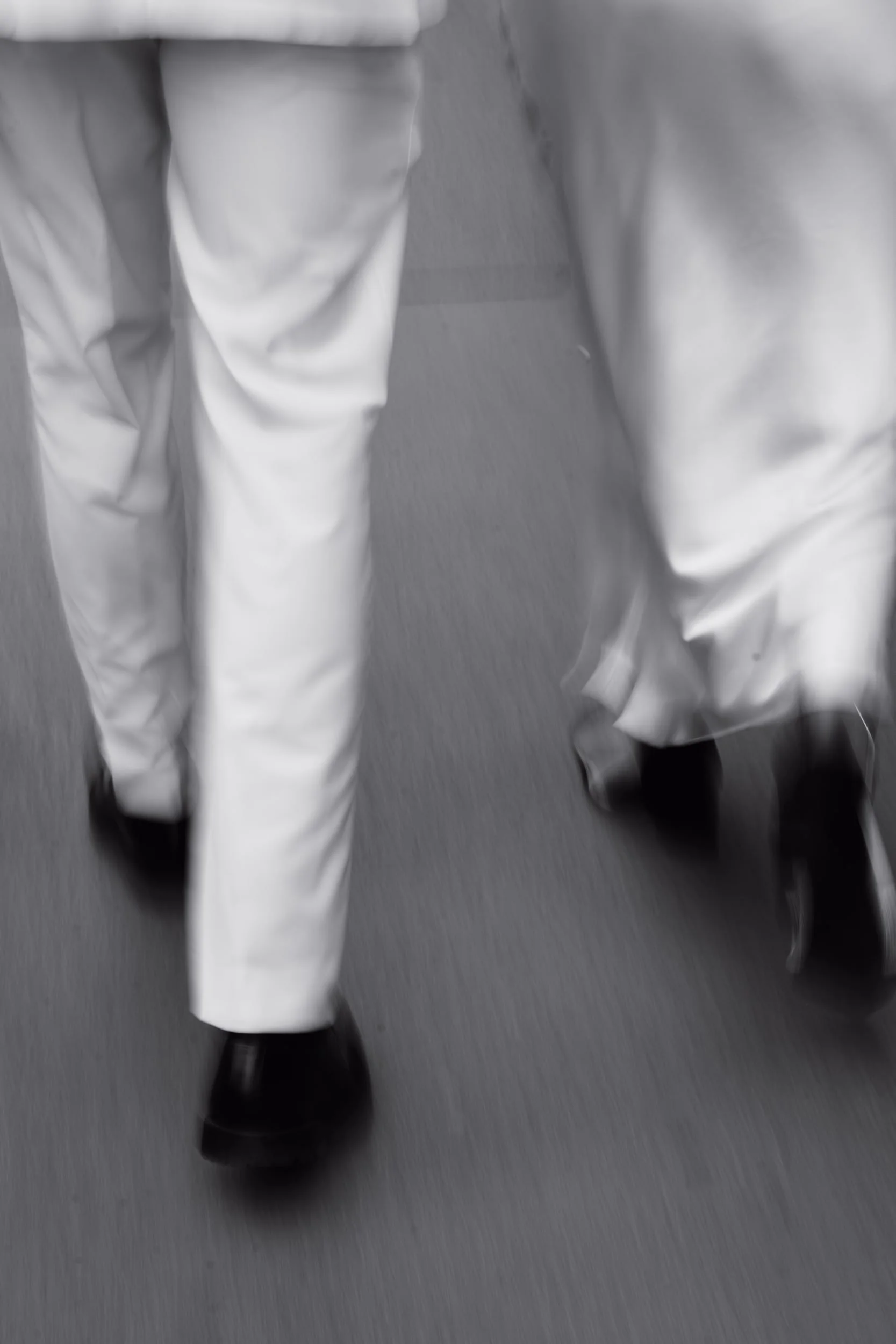 A person wearing white pants and black shoes walking down a gray carpeted hallway.
